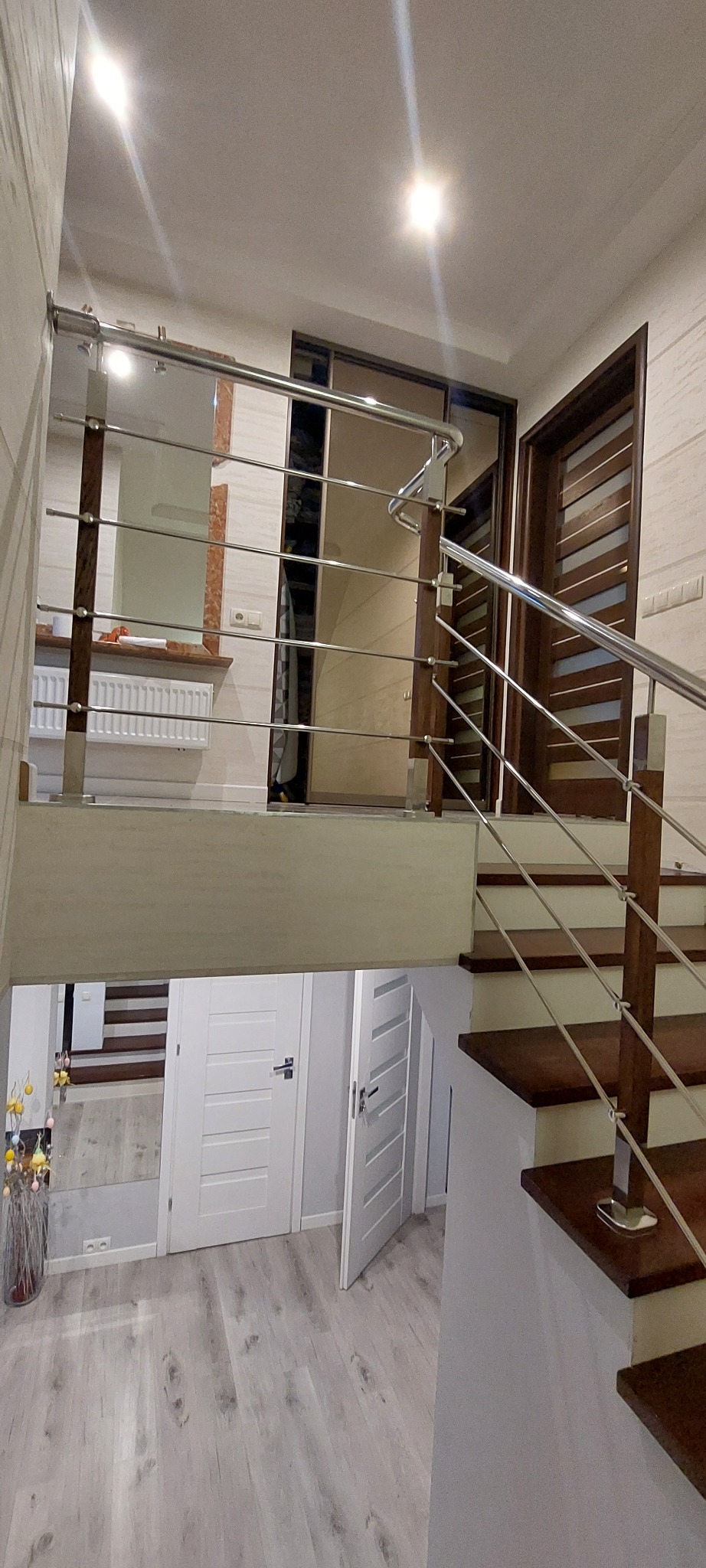 Wnętrze domu z drewnianymi schodami zabezpieczonymi balustradą z elementami drewna i stali nierdzewnej, widok na korytarz z trzema białymi drzwiami.