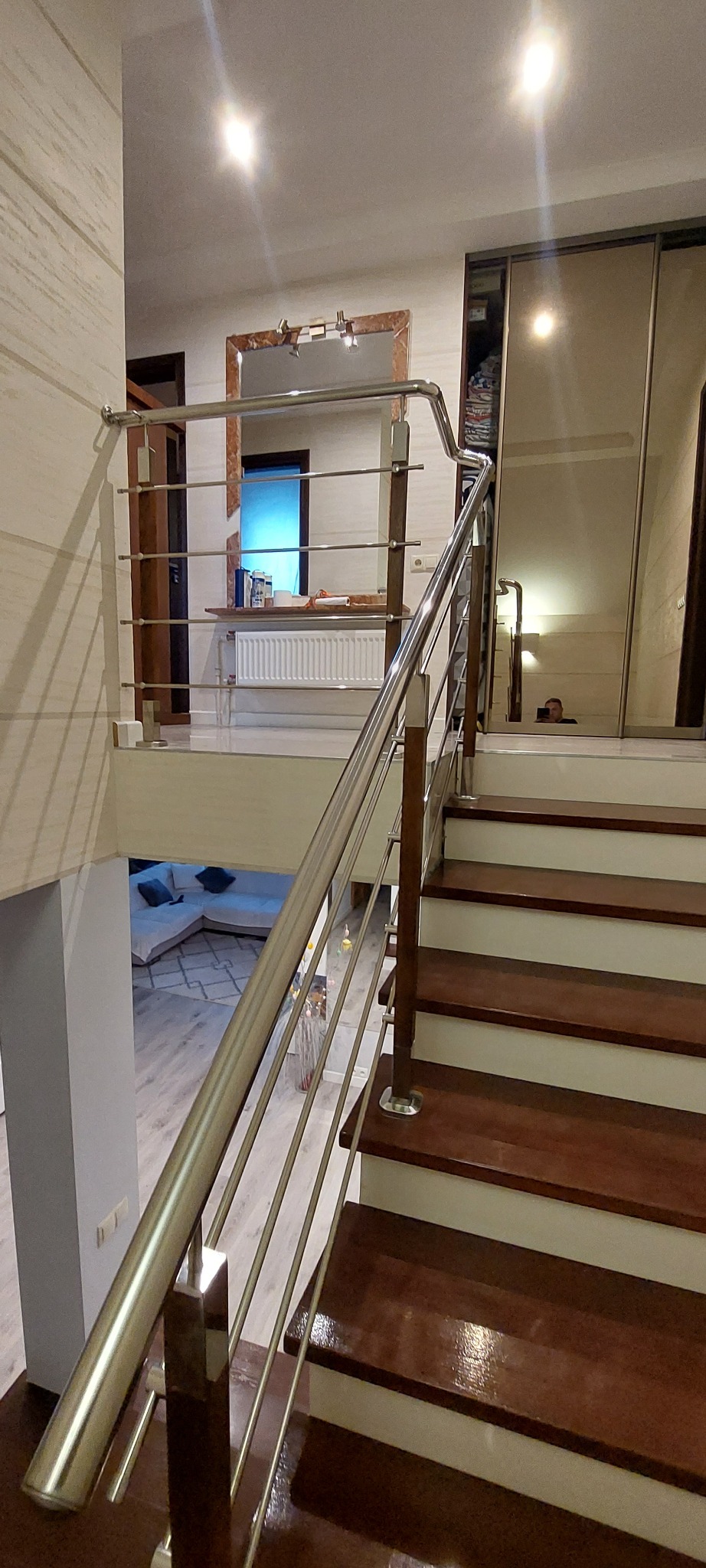 Wnętrze domu z balustradą nierdzewną z drewnianymi słupkami, widok na dół schodów z salonem i odbicie osoby w lustrzanej szafie.