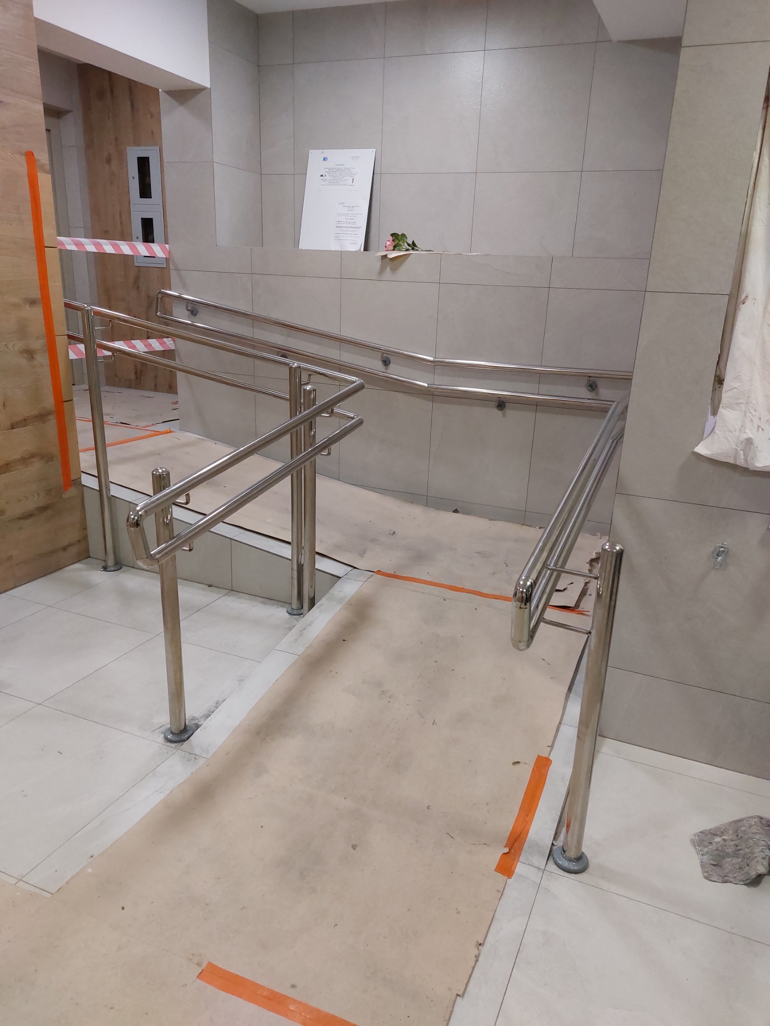 Montaż poręczy ze stali nierdzewnej przy schodach i rampie, zabezpieczone pomarańczową taśmą, widoczne płytki podłogowe i ścienne, w tle dokumentacja techniczna.
