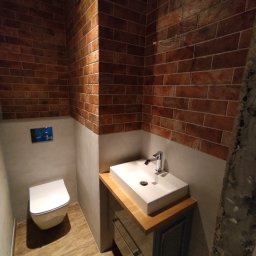 Piotr - Nowoczesna łazienka z ceglaną ścianą, białą umywalką nablatową na drewnianym blacie i podwieszaną toaletą, wykończona w industrialnym stylu.