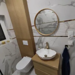 Nowoczesna łazienka z marmurowymi płytkami, okrągłym lustrem w drewnianej ramie, białą umywalką z złotymi akcentami, szafką z jasnego drewna i wiszącą toaletą z przyciskiem spłukiwania.