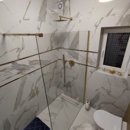 Piotr - Nowoczesna łazienka z kabiną prysznicową typu walk-in, wykończona białymi płytkami imitującymi marmur z akcentami złota, ciemnoniebieski dywanik łazienkowy i dekoracje na parapecie okiennym.