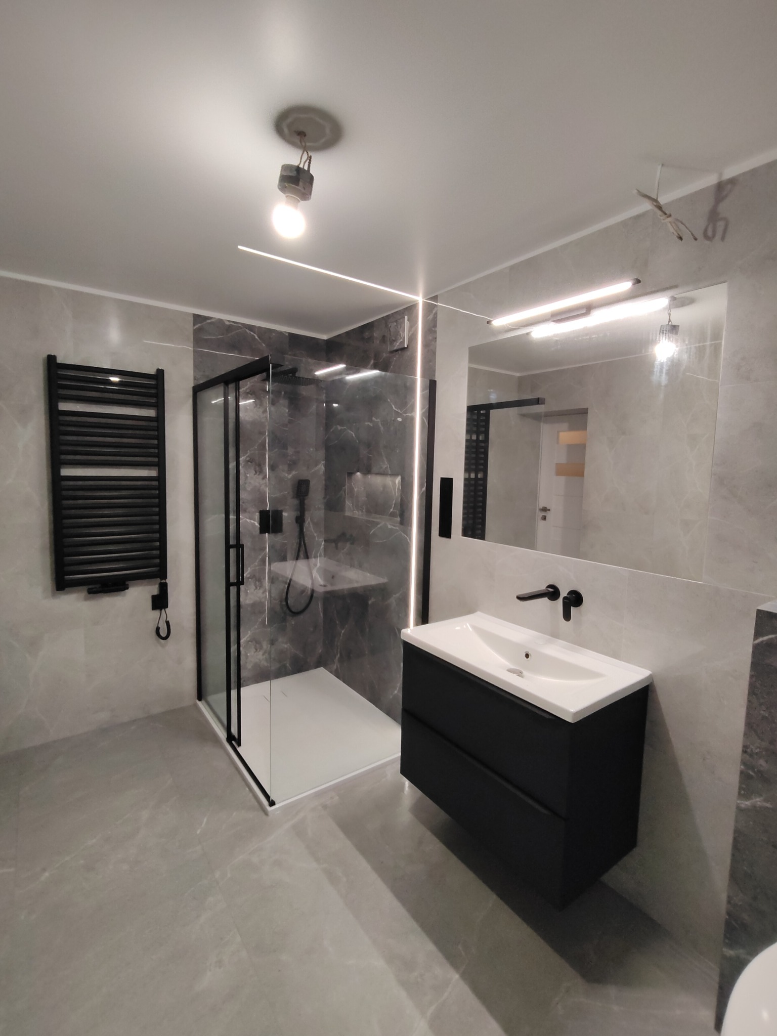 Nowoczesna łazienka w szarościach z czarnymi akcentami: kabina prysznicowa typu walk-in, grzejnik drabinkowy, szafka pod umywalkę z czarnymi frontami i baterią, duże lustro z oświetleniem LED.