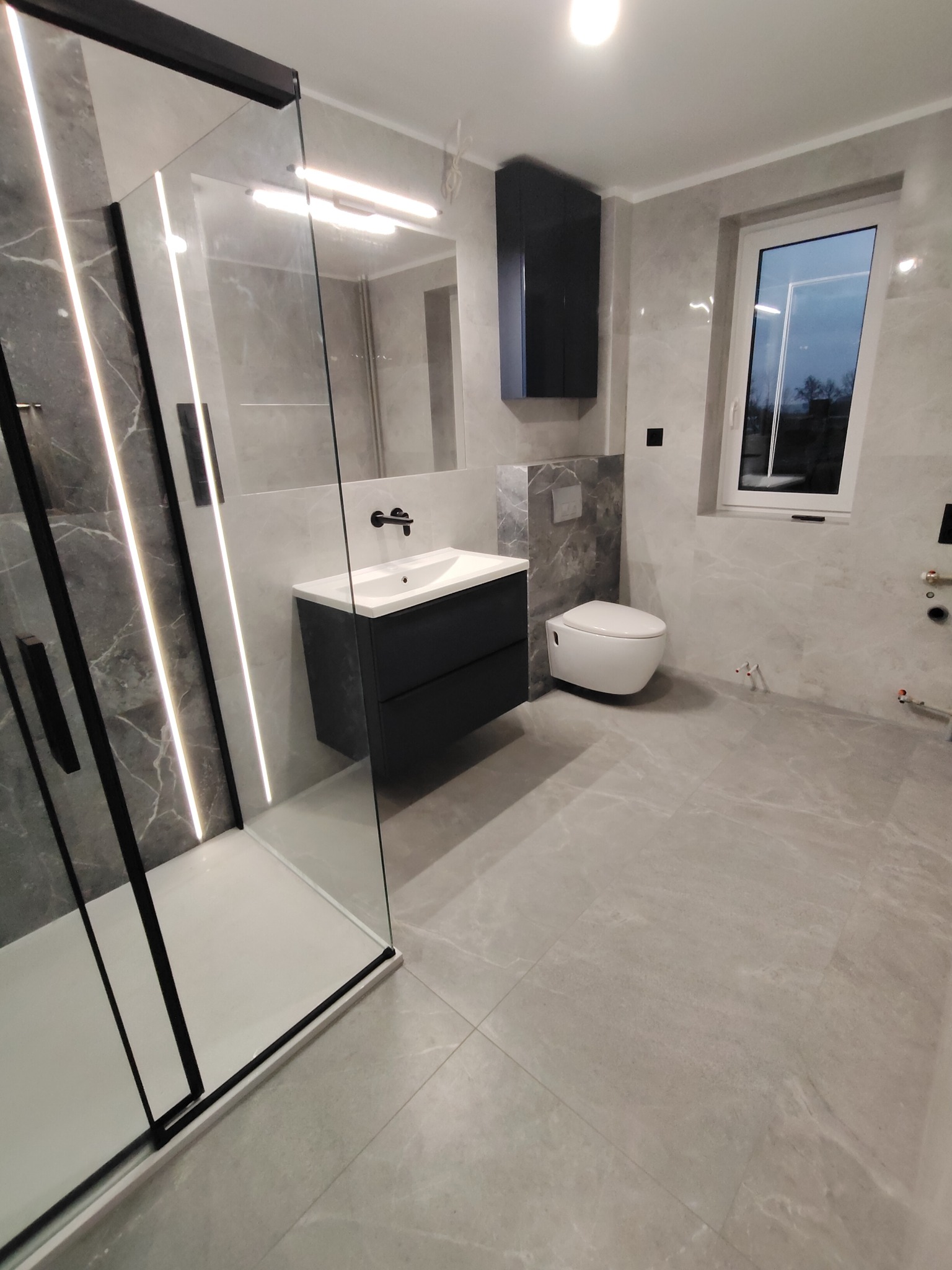 Nowoczesna łazienka wykończona w szarościach, z kabiną prysznicową typu walk-in z czarnymi profilami, wiszącą szafką nad umywalką z czarną armaturą oraz podwieszaną toaletą.