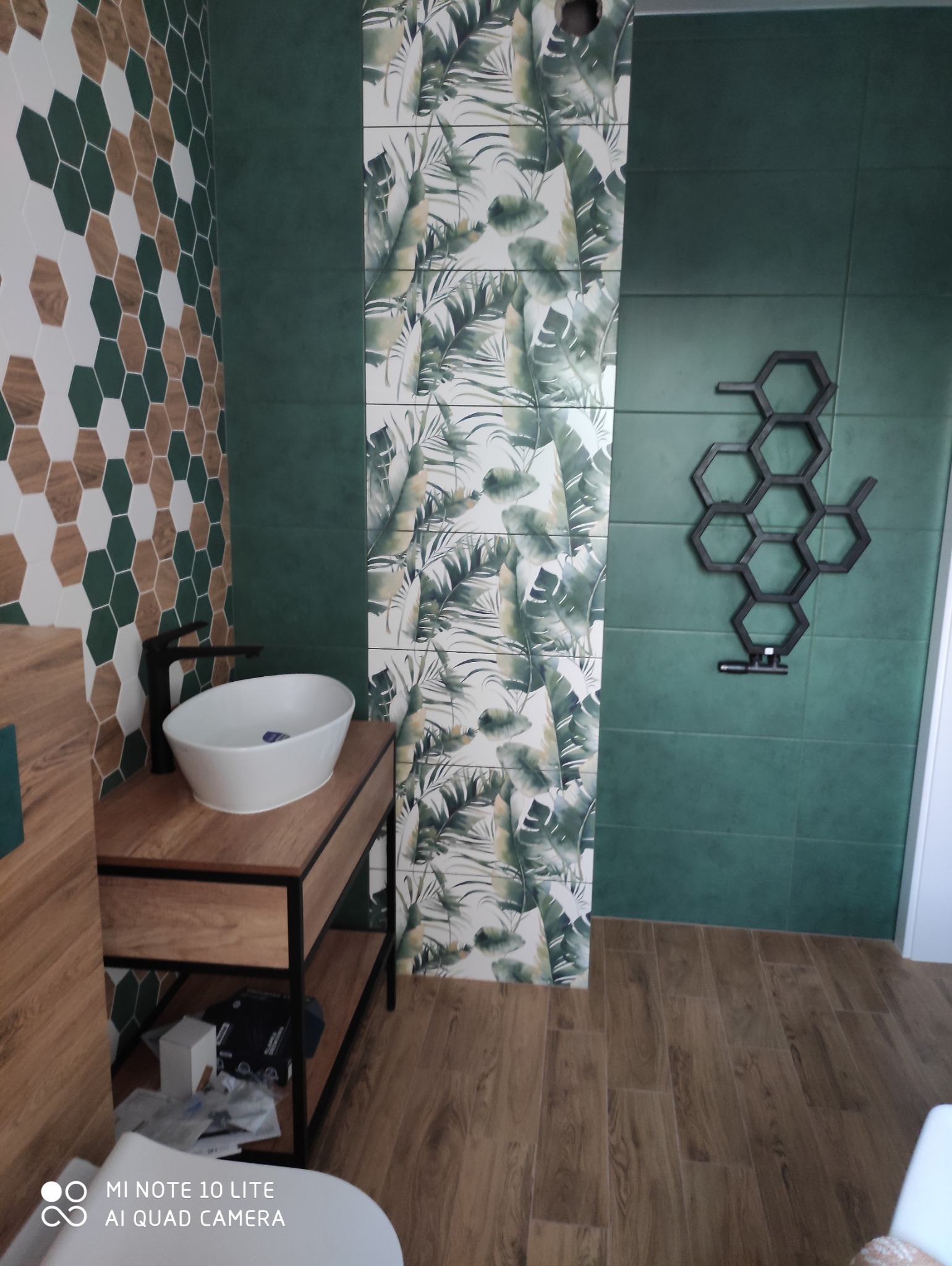 Nowoczesna łazienka z zielonymi płytkami, heksagonalnymi wzorami i tropikalnym motywem na ścianie, umywalka nablatowa na drewnianej szafce z czarnym kranem i grzejnik drabinkowy w kształcie plastra...