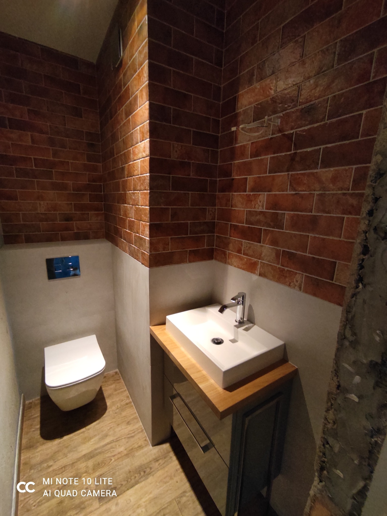 Nowoczesna łazienka z ceglaną ścianą, białą umywalką nablatową na drewnianym blacie i podwieszaną toaletą, wykończona w industrialnym stylu.