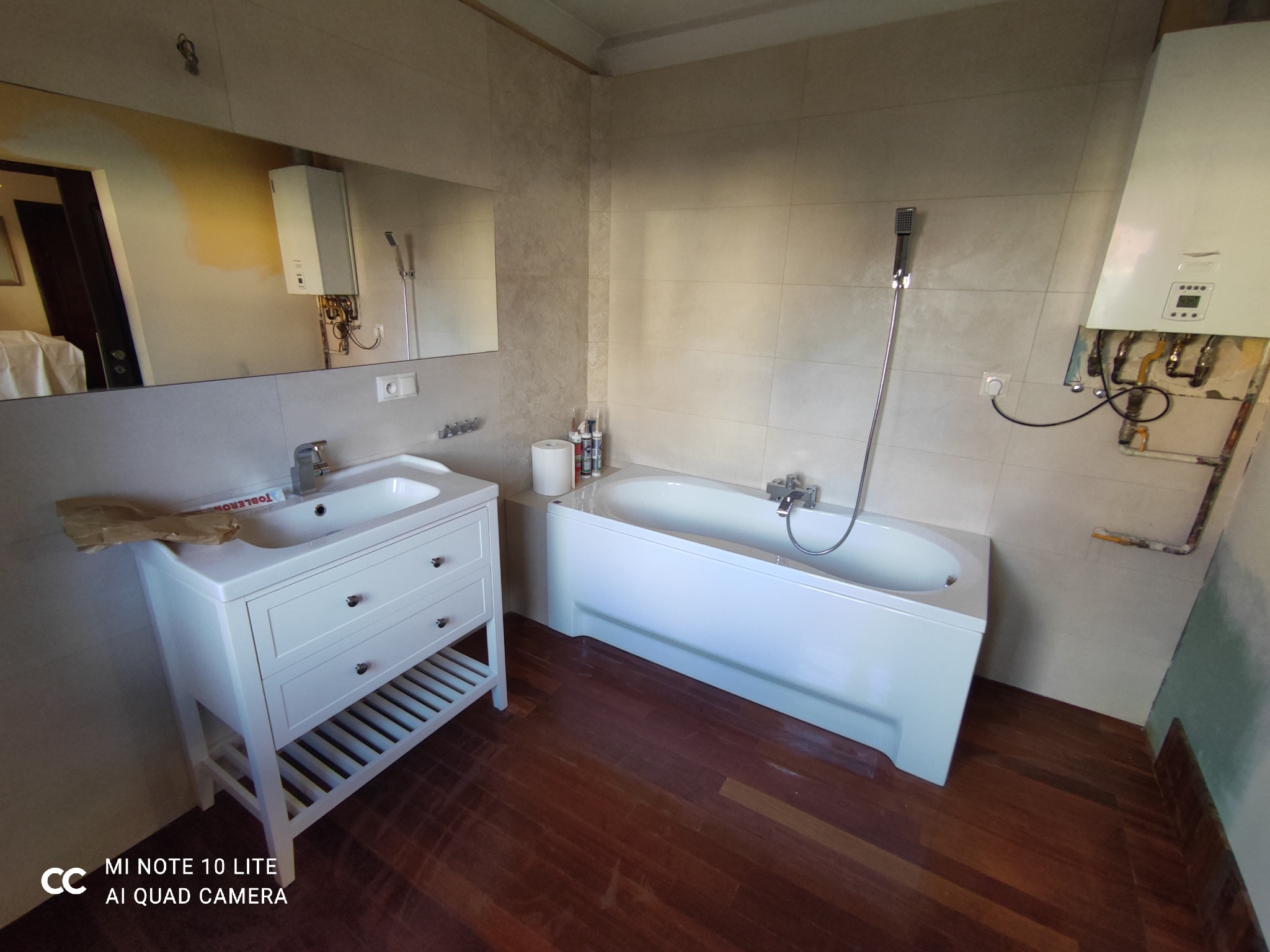 Biała łazienka z wanną, umywalką z szafką i brązową podłogą. Na ścianie widoczny bojler z odkrytymi rurami i niedokończone płytki. Na umywalce leży opakowanie czekolady.