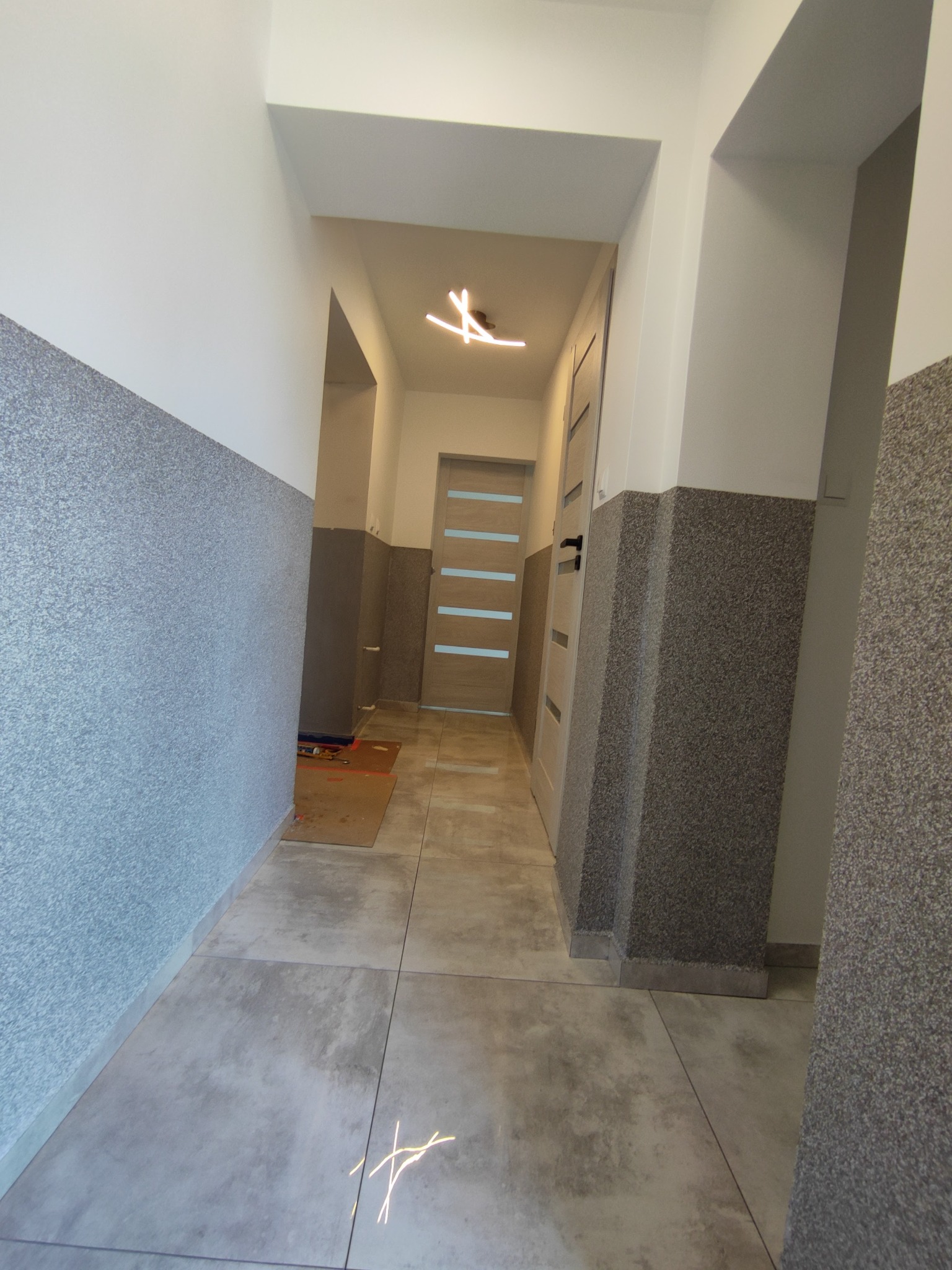 Korytarz w trakcie remontu: położone płytki podłogowe, ściany częściowo pokryte szarą strukturą, widoczne narzędzia i materiały budowlane w głębi.