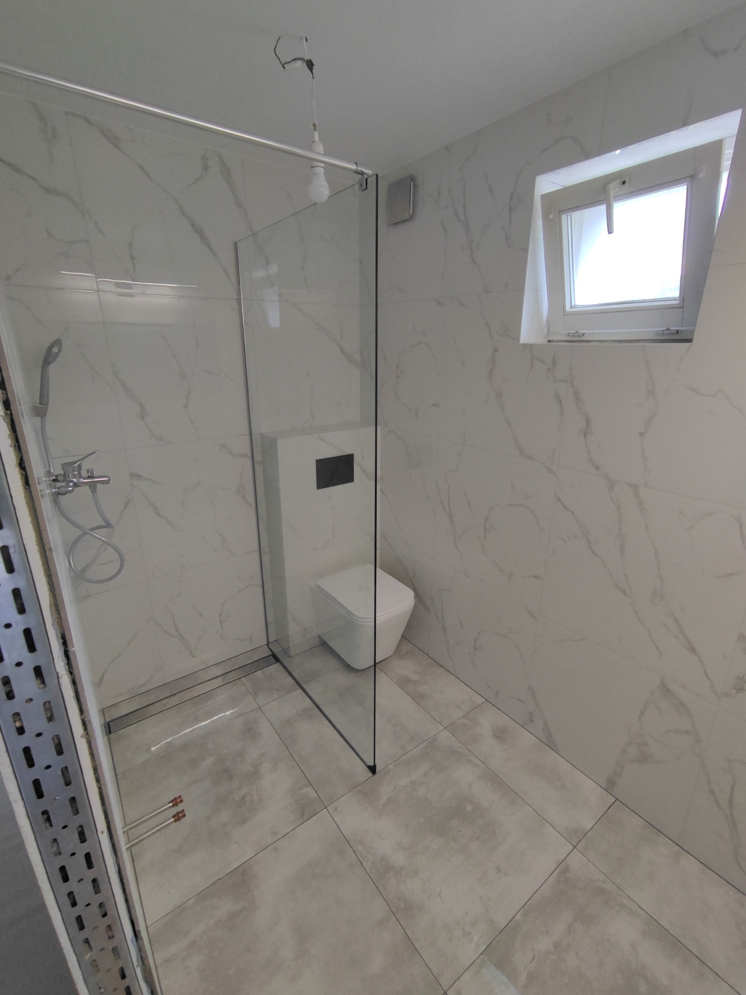 Nowoczesna łazienka z kabiną prysznicową typu walk-in, podwieszaną toaletą i płytkami imitującymi marmur na ścianach; widoczne rury wystające z podłogi przed wykończeniem.