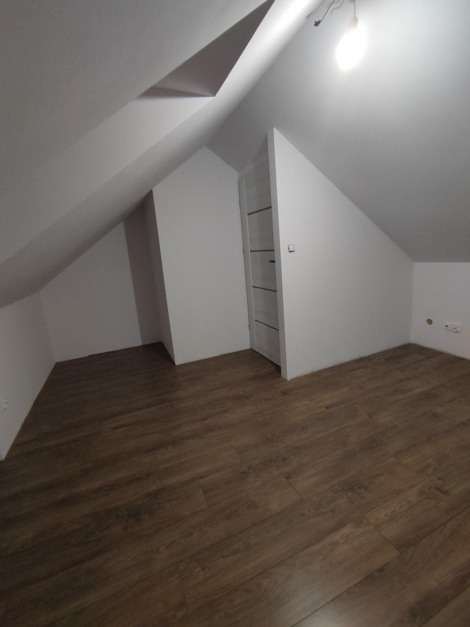 Poddasze z drewnianą podłogą, białe ściany i zabudowana szafa wnękowa z białymi drzwiami, widoczna żarówka.