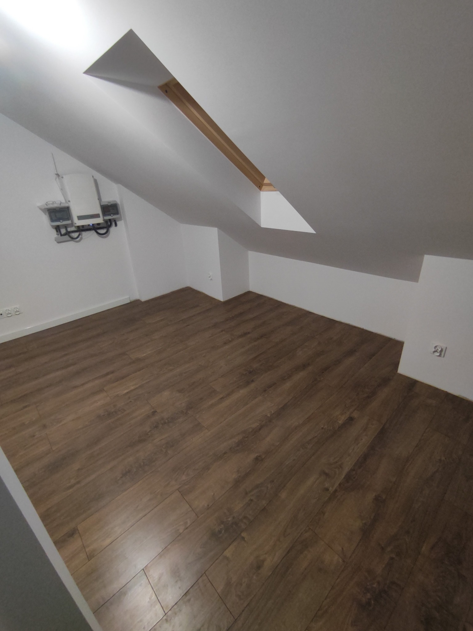 Poddasze z drewnopodobną podłogą, skośnym sufitem z oknem dachowym i białymi ścianami, na których widać instalację elektryczną.