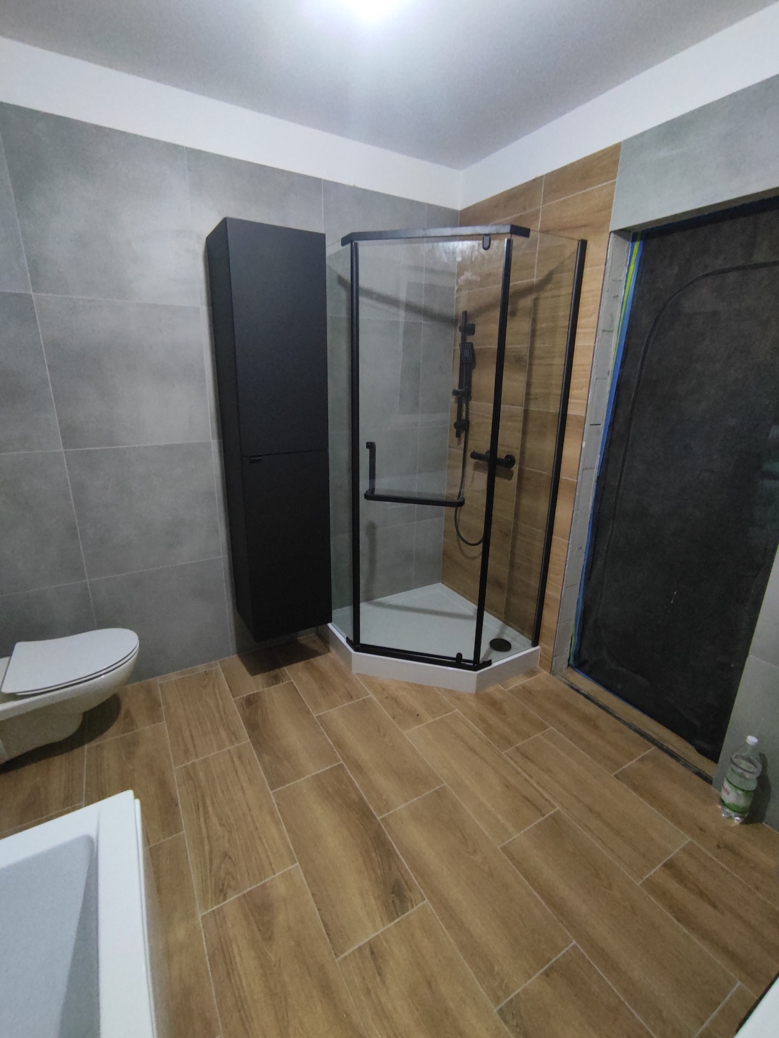 Nowoczesna łazienka po remoncie: kabina prysznicowa z czarnymi profilami, wisząca szafka w kolorze czarnym, płytki imitujące drewno na podłodze, szare płytki na ścianach, częściowo wykończone...