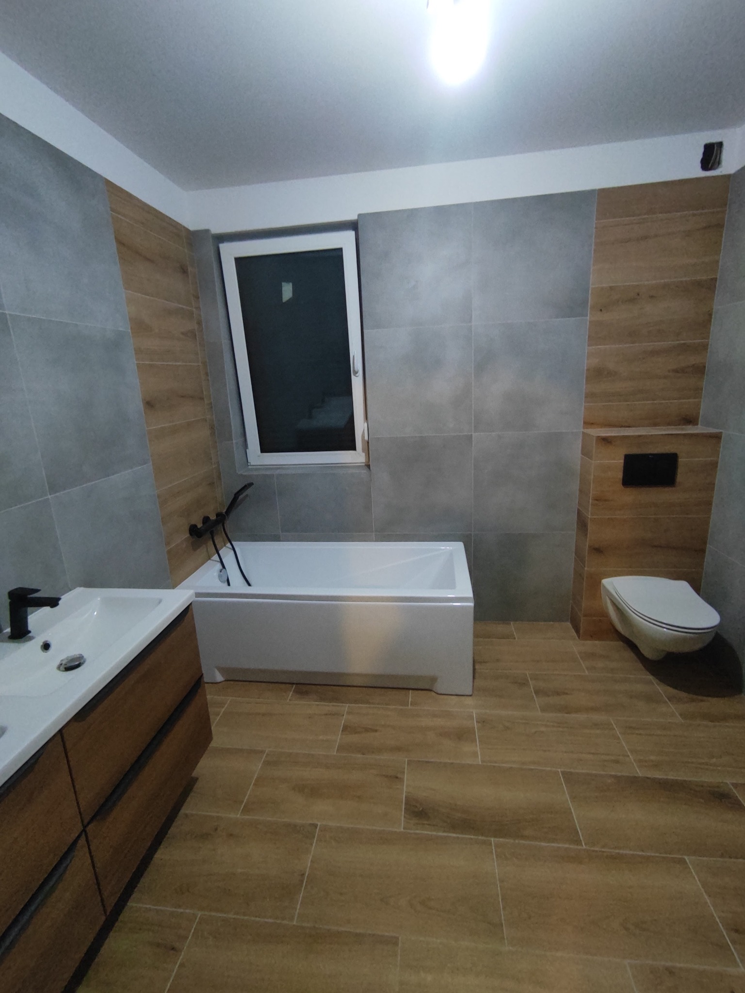 Nowoczesna łazienka z płytkami imitującymi drewno na podłodze i częściowo na ścianach, kontrastujące z szarymi płytkami ściennymi, biała wanna i umywalka z brązową szafką, minimalistyczna toaleta...