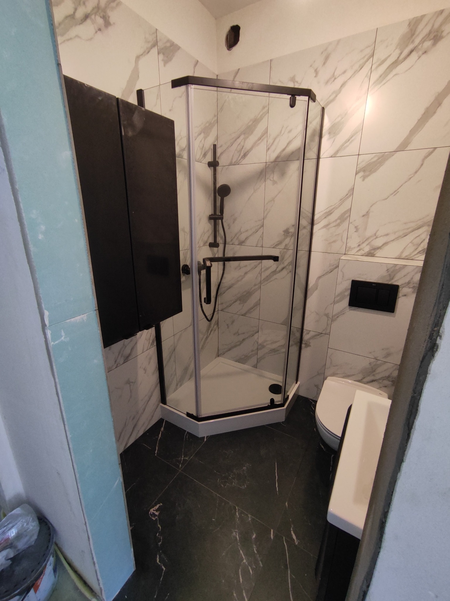 Nowoczesna łazienka z kabiną prysznicową typu walk-in z czarnymi elementami, marmurowymi płytkami na ścianach i ciemną podłogą, częściowo widoczna szafka i toaleta.