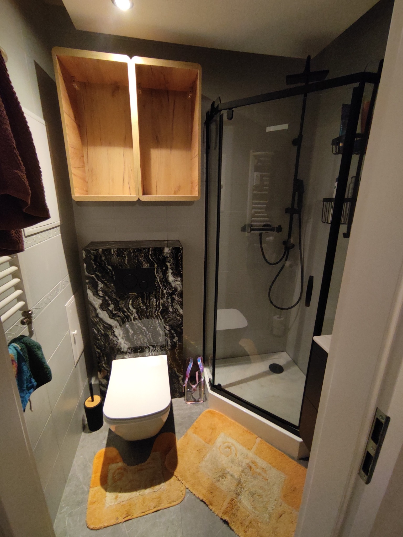 Nowoczesna łazienka z kabiną prysznicową z czarnymi ramami, podwieszaną toaletą z czarnym marmurowym panelem, drewnianymi szafkami bez frontów i pomarańczowymi dywanikami.