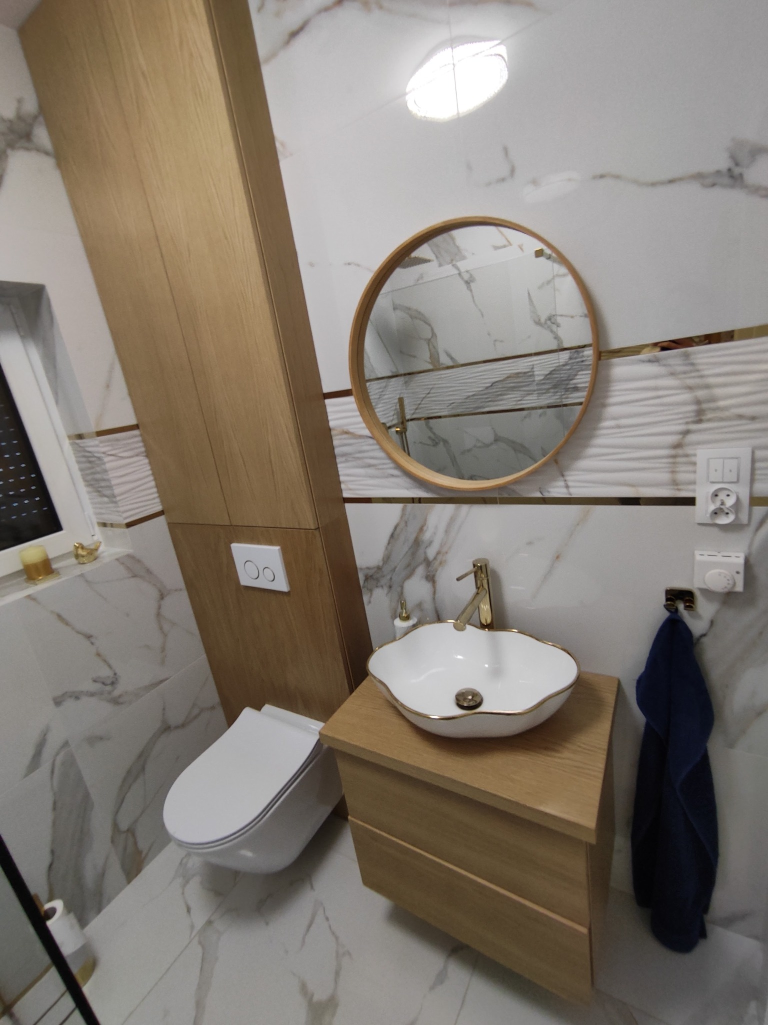 Nowoczesna łazienka z marmurowymi płytkami, okrągłym lustrem w drewnianej ramie, białą umywalką z złotymi akcentami, szafką z jasnego drewna i wiszącą toaletą z przyciskiem spłukiwania.