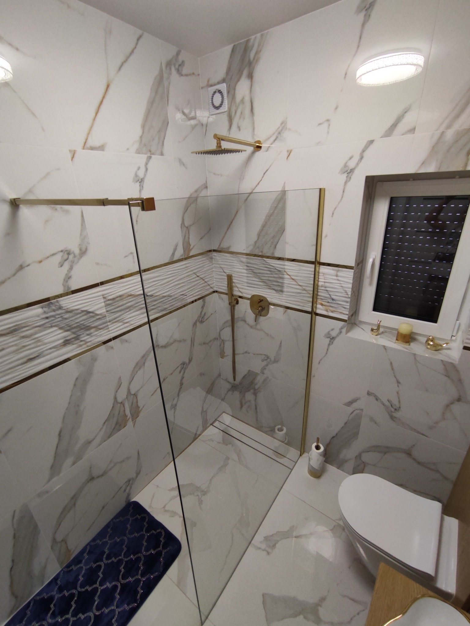 Nowoczesna łazienka z kabiną prysznicową typu walk-in, wykończona białymi płytkami imitującymi marmur z akcentami złota, ciemnoniebieski dywanik łazienkowy i dekoracje na parapecie okiennym.