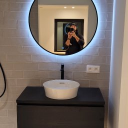 Elektro System - Nowoczesna łazienka z okrągłym lustrem LED, umywalką nablatową i szarą glazurą, widoczne gniazdka elektryczne.