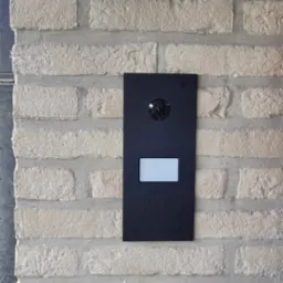 Czarny panel wideodomofonu z kamerą i wyświetlaczem zamontowany na ścianie z jasnej cegły klinkierowej.