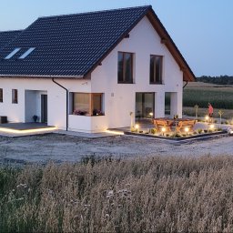 Elektro System - Nowoczesny dom jednorodzinny z podświetlonym tarasem i ogrodem w wieczornym oświetleniu, widoczne lampy ogrodowe, drewniany stół z ławkami i osoba w oddali.