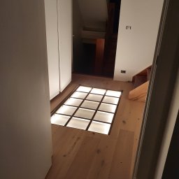 Elektro System - Podświetlana szklana podłoga w korytarzu z drewnianą podłogą i białymi szafami w tle.