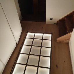 Elektro System - Podświetlana podłoga z kwadratowymi panelami wkomponowana w drewnianą podłogę, widok z góry na korytarz z elementami w trakcie wykończenia.