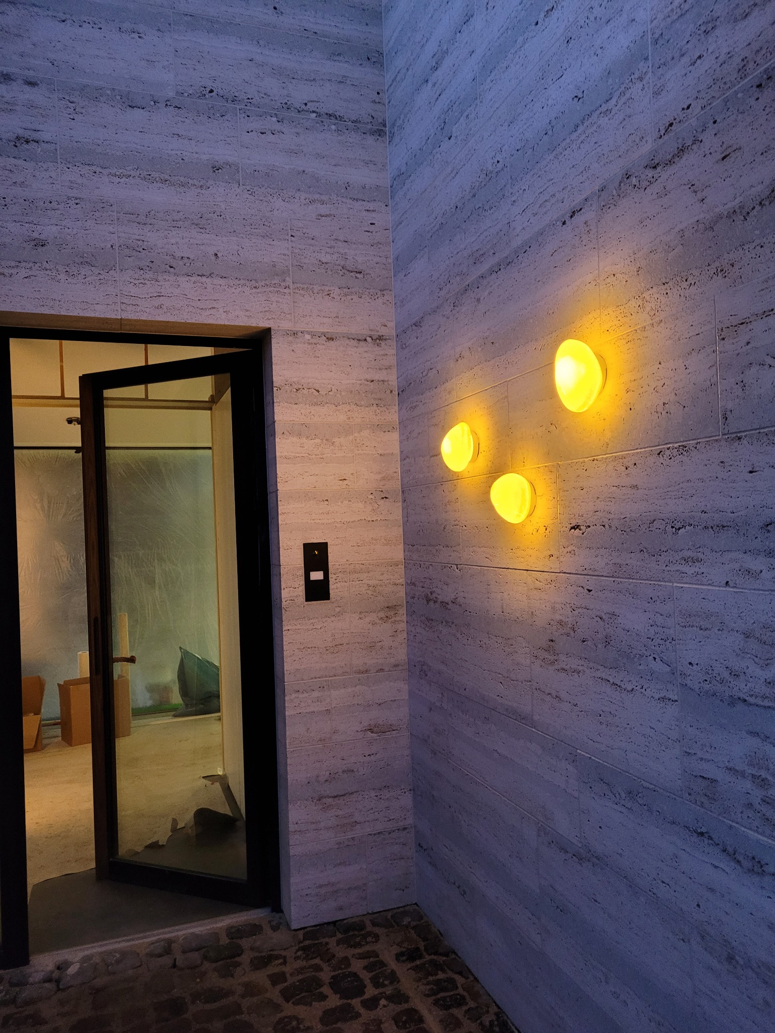 Ściana budynku wyłożona kamieniem trawertynowym z trzema okrągłymi lampami ściennymi o ciepłej barwie światła, obok otwarte, szklane drzwi wejściowe z czarną ramą, w tle widoczne wnętrze budynku.