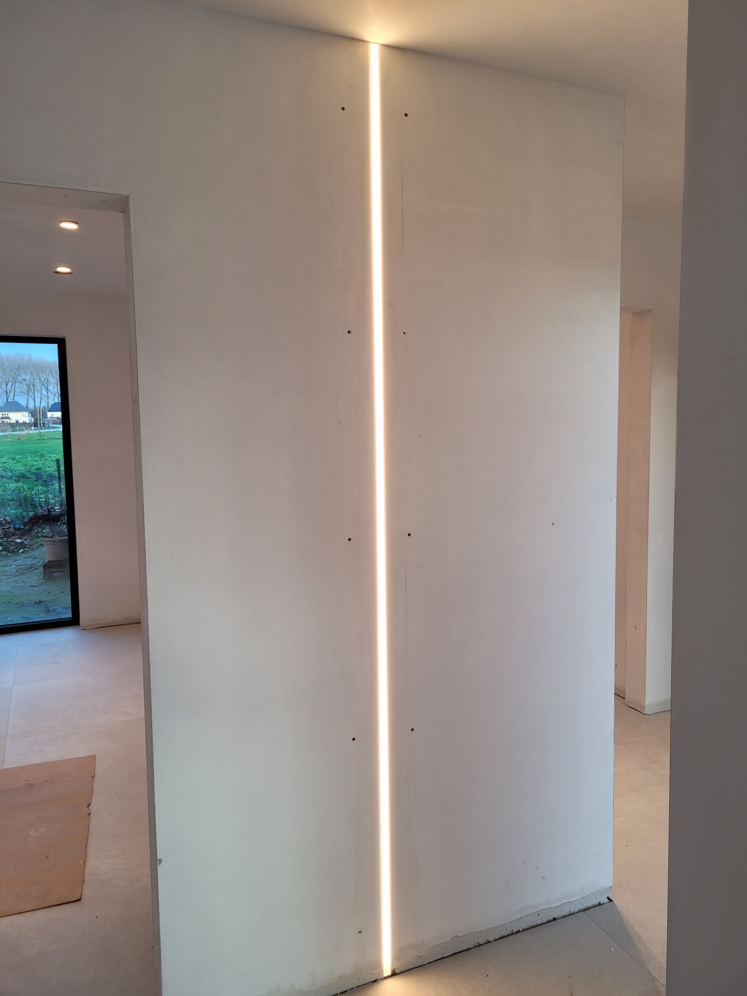 Wąska pionowa szczelina światła w ścianie z gładzią, widoczne niedoskonałości wykończenia, surowy stan wnętrza z widokiem na okno i krajobraz za nim.