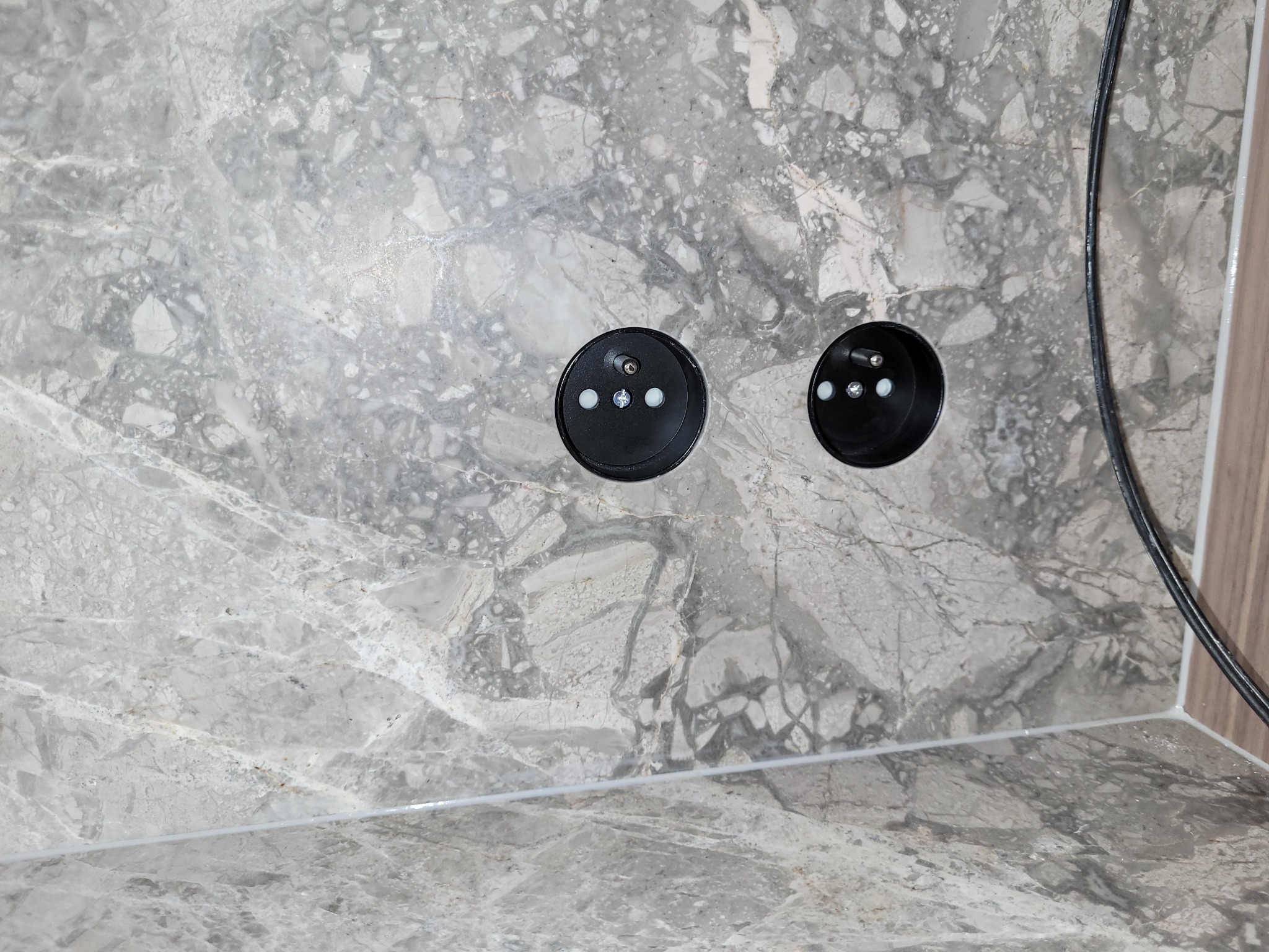 Dwa czarne gniazdka elektryczne zamontowane w marmurowej ścianie, widoczny przewód elektryczny po prawej stronie.