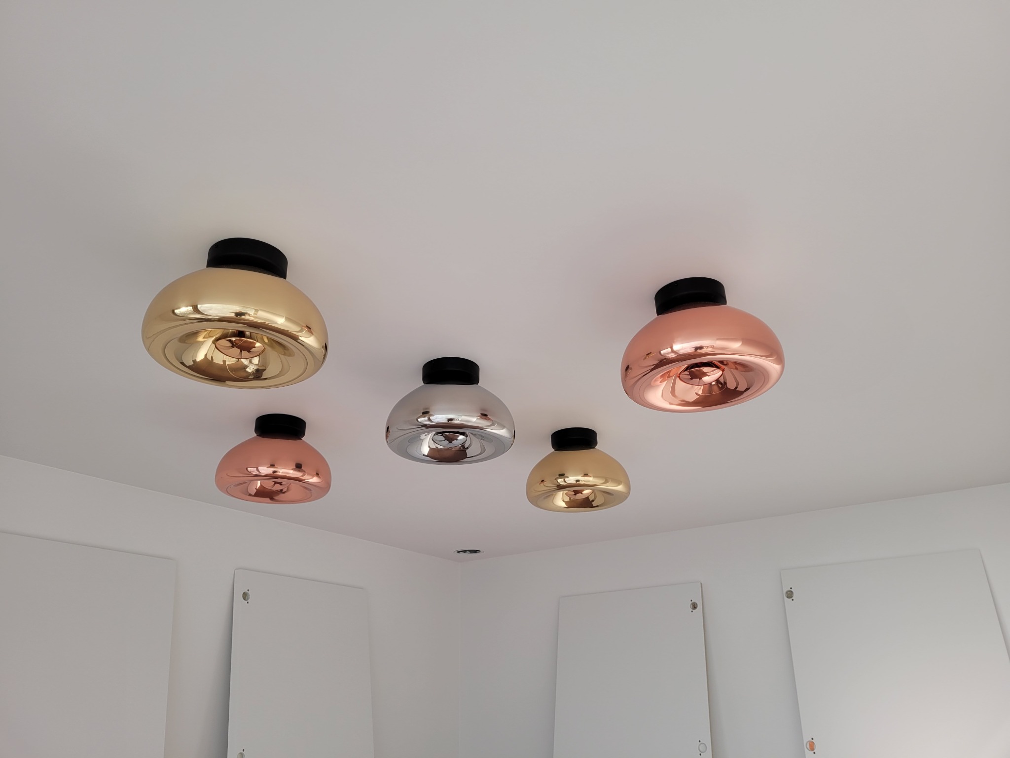 Sufitowe lampy w odcieniach złota, srebra i miedzi, rozmieszczone asymetrycznie na białym suficie w nowoczesnym wnętrzu.