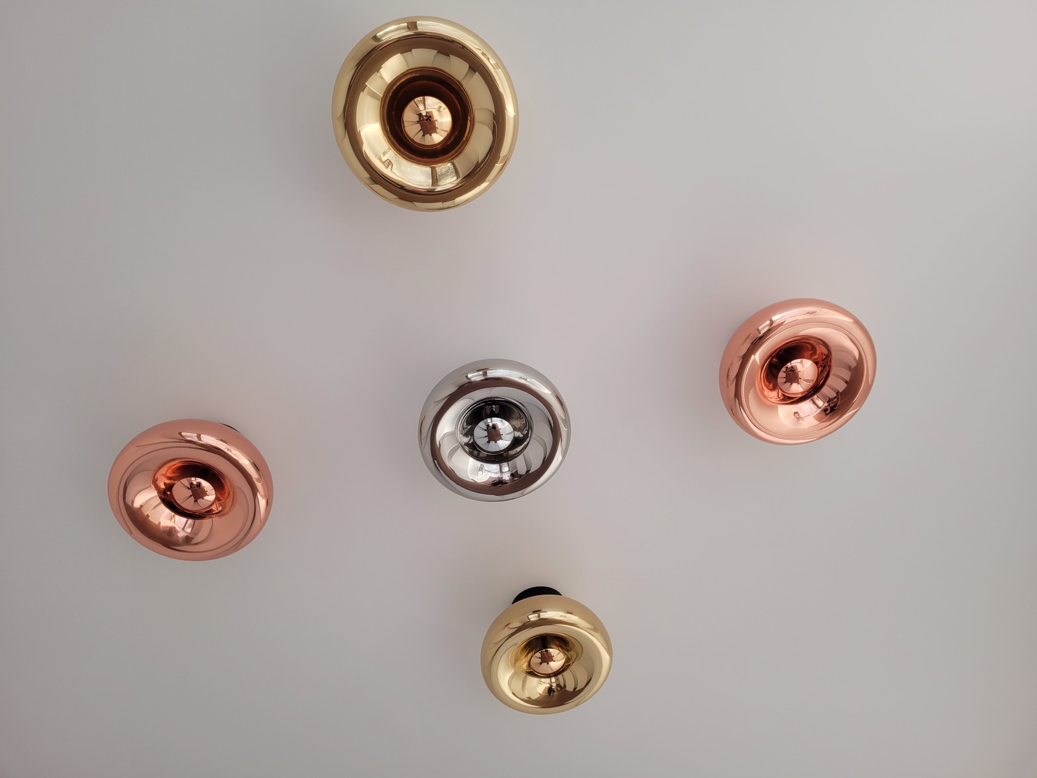 Pięć okrągłych lamp sufitowych w kolorach miedzi, złota i srebra, rozmieszczonych asymetrycznie na białym suficie, z widocznymi odbiciami światła w metalicznych obudowach.