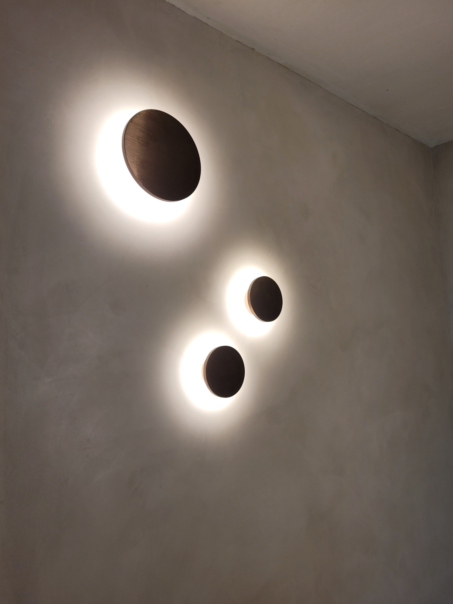 Trzy okrągłe lampy ścienne z brązowymi elementami, emitujące ciepłe światło na szarej, teksturowanej ścianie.