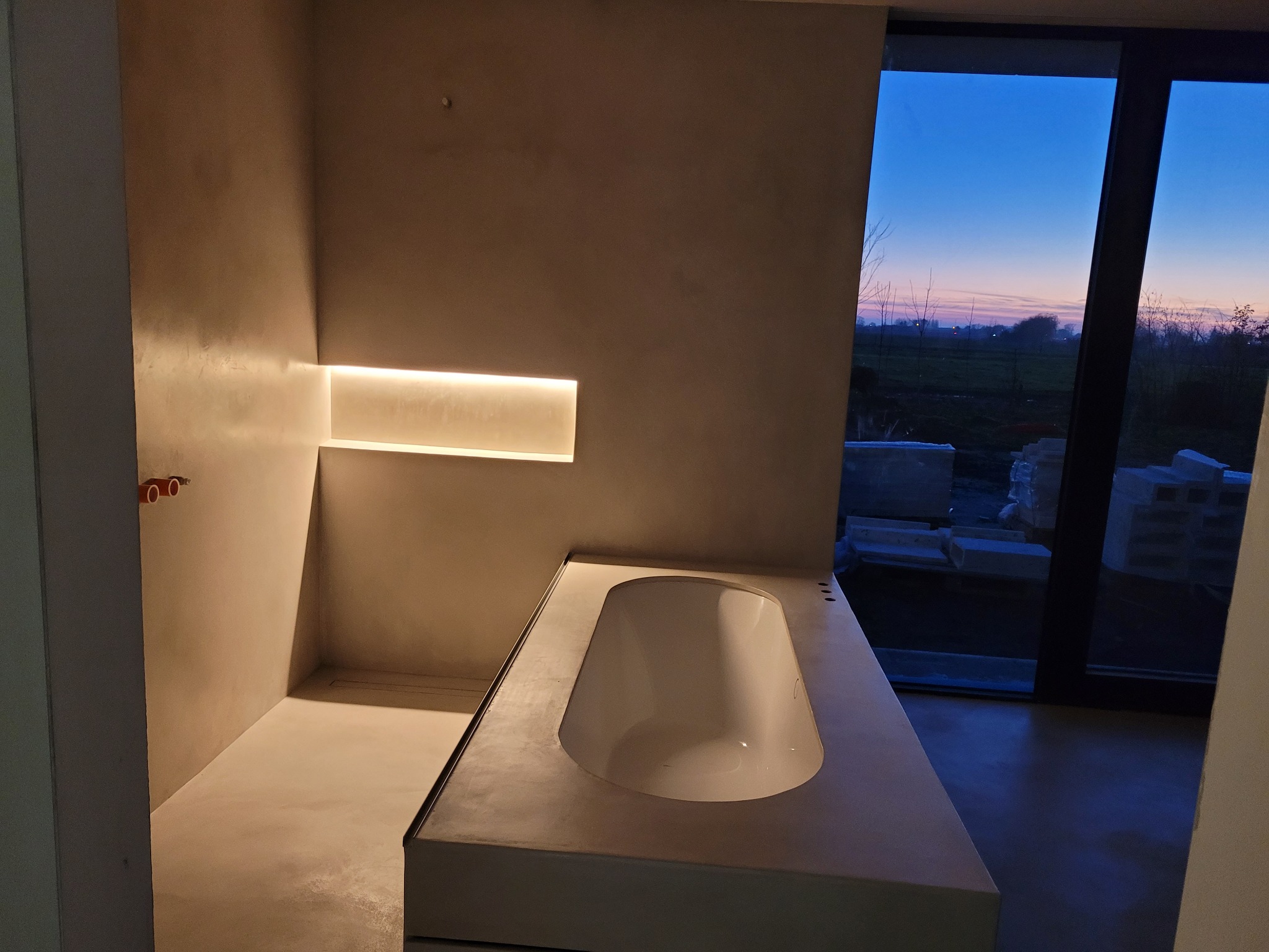 Minimalistyczna łazienka w surowym stylu z wbudowaną wanną obłożoną betonem architektonicznym, podświetlona listwą LED i z widokiem na zachodzące słońce za oknem.