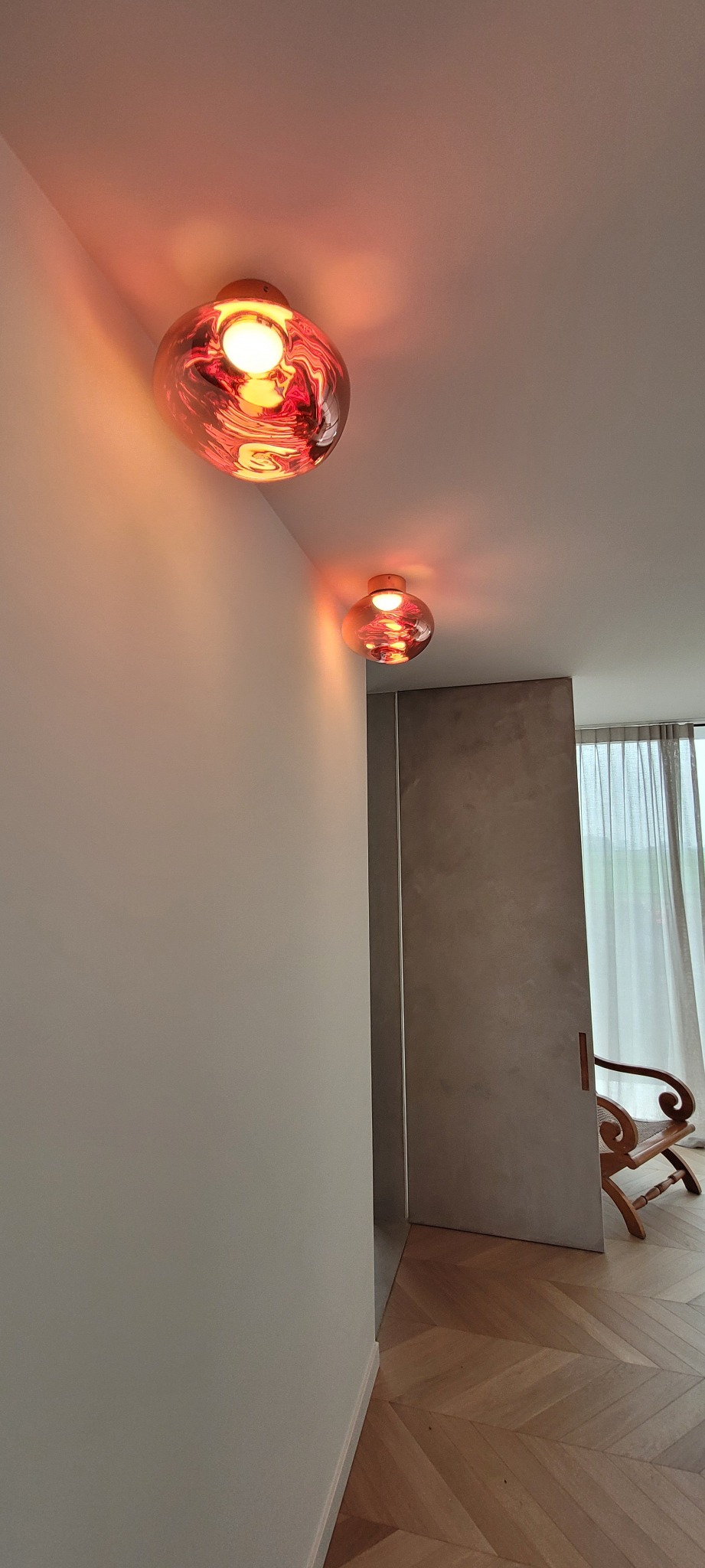 Dwie szklane lampy sufitowe w kolorze czerwonym, o nieregularnym kształcie, oświetlają białą ścianę i fragment parkietu w nowoczesnym wnętrzu.