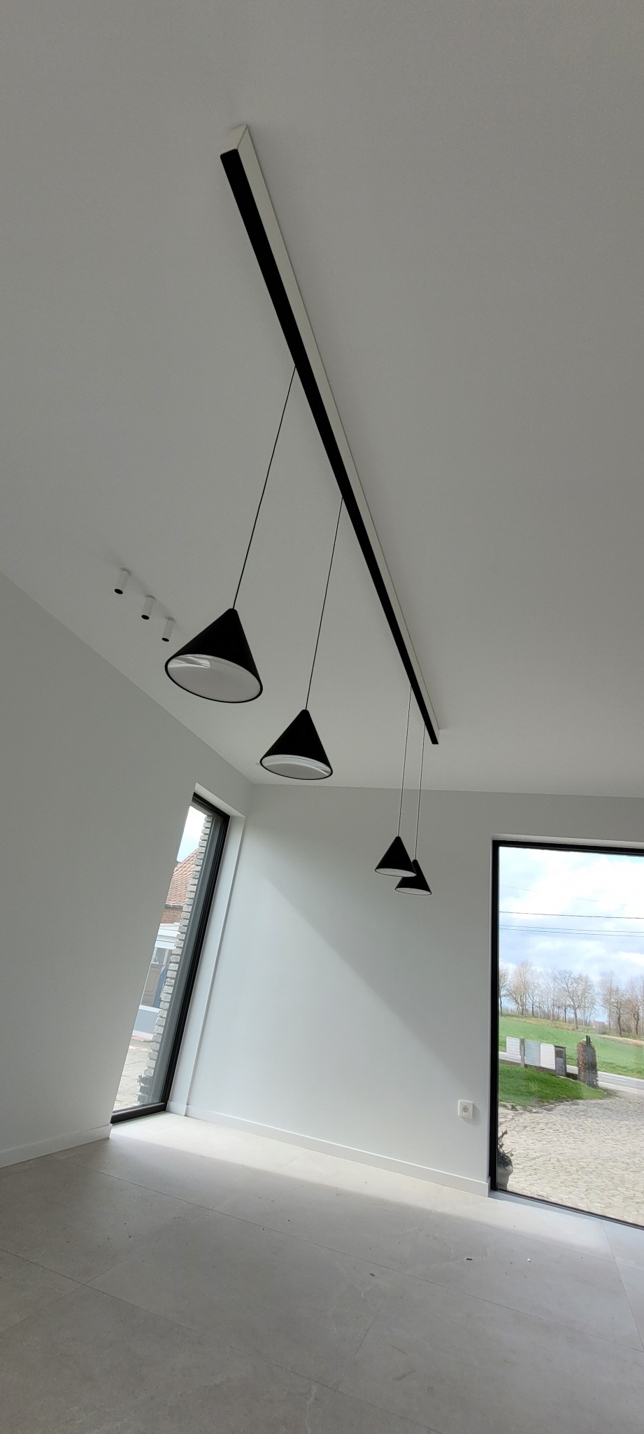 Nowoczesne oświetlenie sufitowe z czterema czarnymi, stożkowymi lampami wiszącymi na cienkich przewodach, zamocowane na długiej, prostokątnej listwie w minimalistycznym wnętrzu z dużymi oknami.