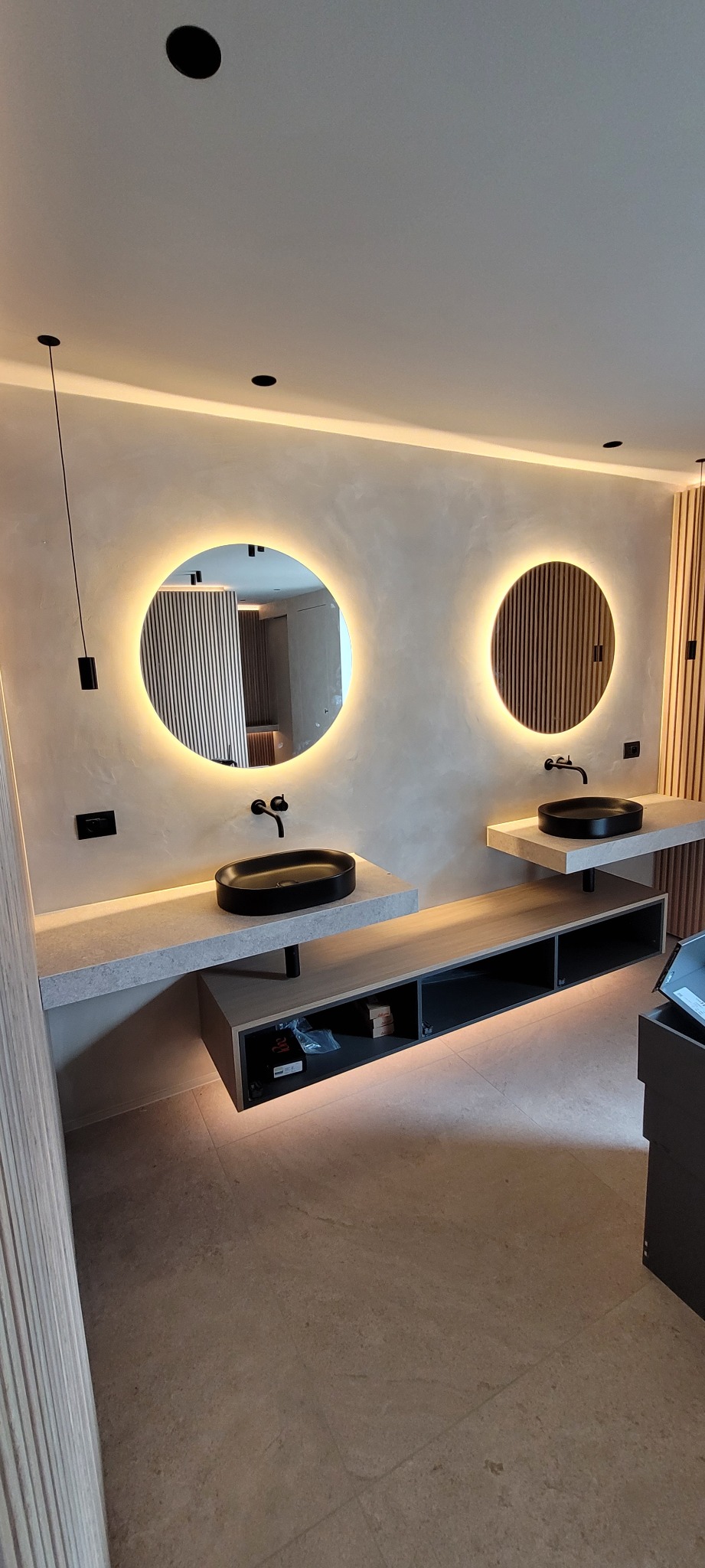 Nowoczesna łazienka z dwoma okrągłymi lustrami podświetlanymi LED, czarnymi umywalkami nablatowymi i minimalistyczną armaturą, oświetleniem punktowym oraz podświetlaną szafką podwieszaną.