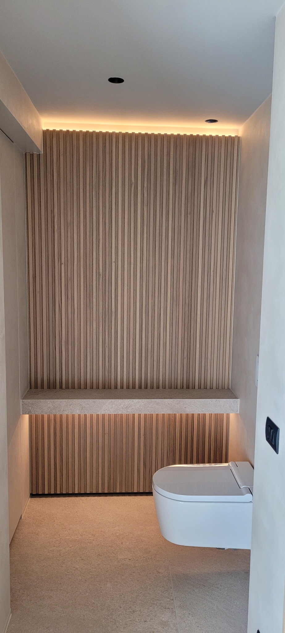 Nowoczesna toaleta z podświetlaną drewnianą ścianą z listew i półką z kamienia, minimalistyczny design.