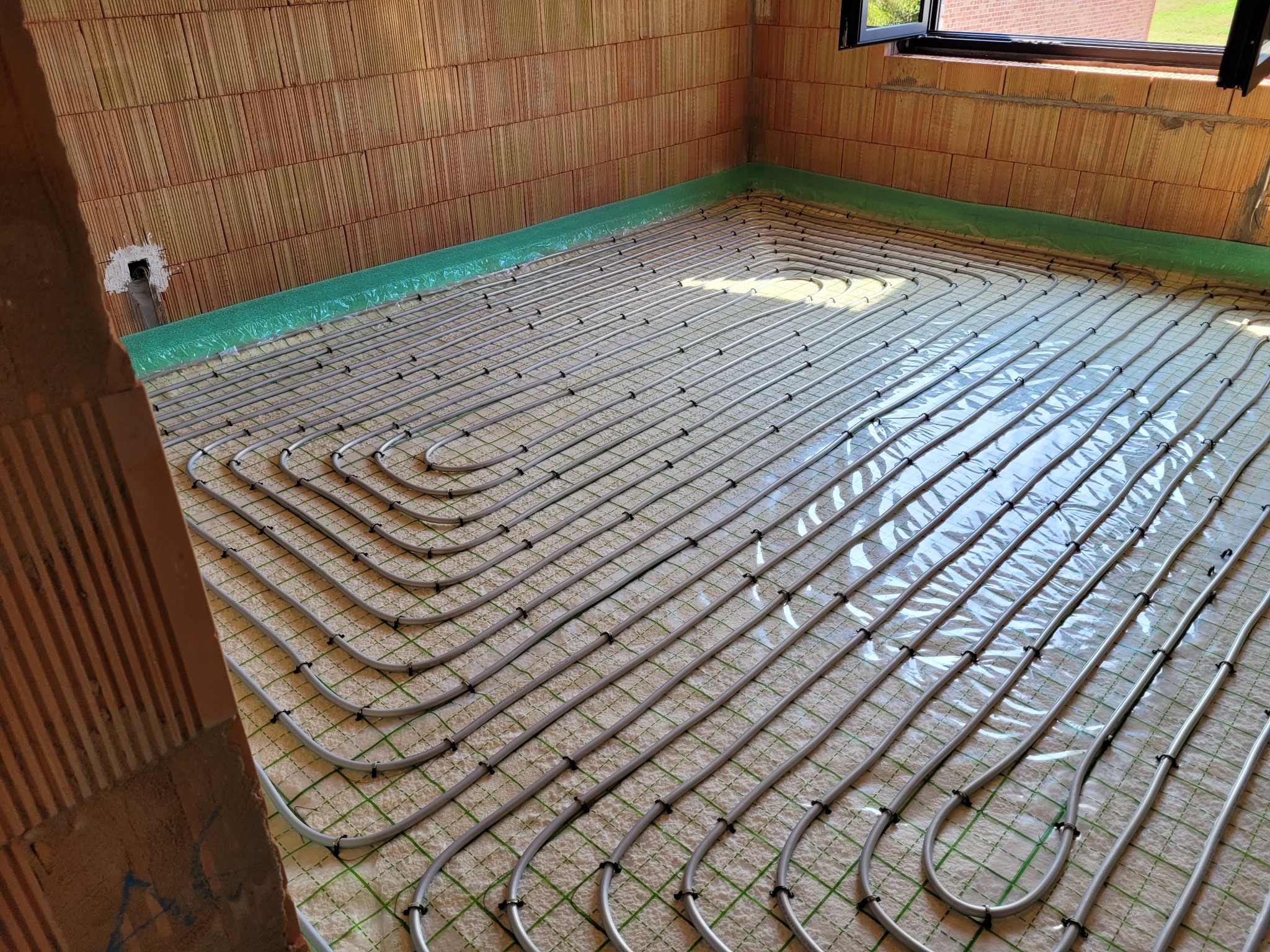 Rozłożone rury ogrzewania podłogowego na izolacji z siatką, widoczne mocowania rur, ściany z pustaków ceramicznych i fragment okna.
