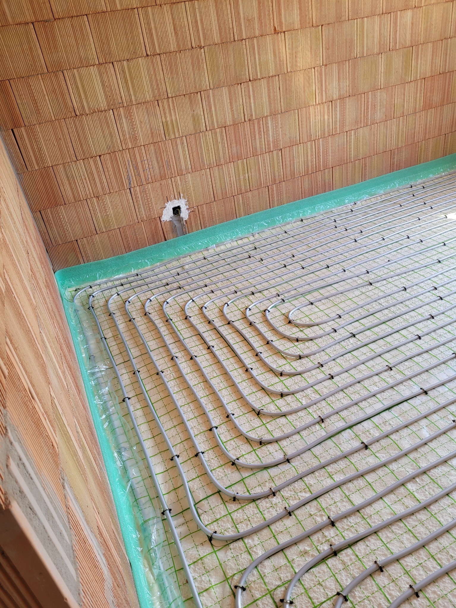 Rozłożona instalacja ogrzewania podłogowego na warstwie izolacyjnej, widoczne rury grzewcze mocowane spinkami, ściany z cegły ceramicznej.