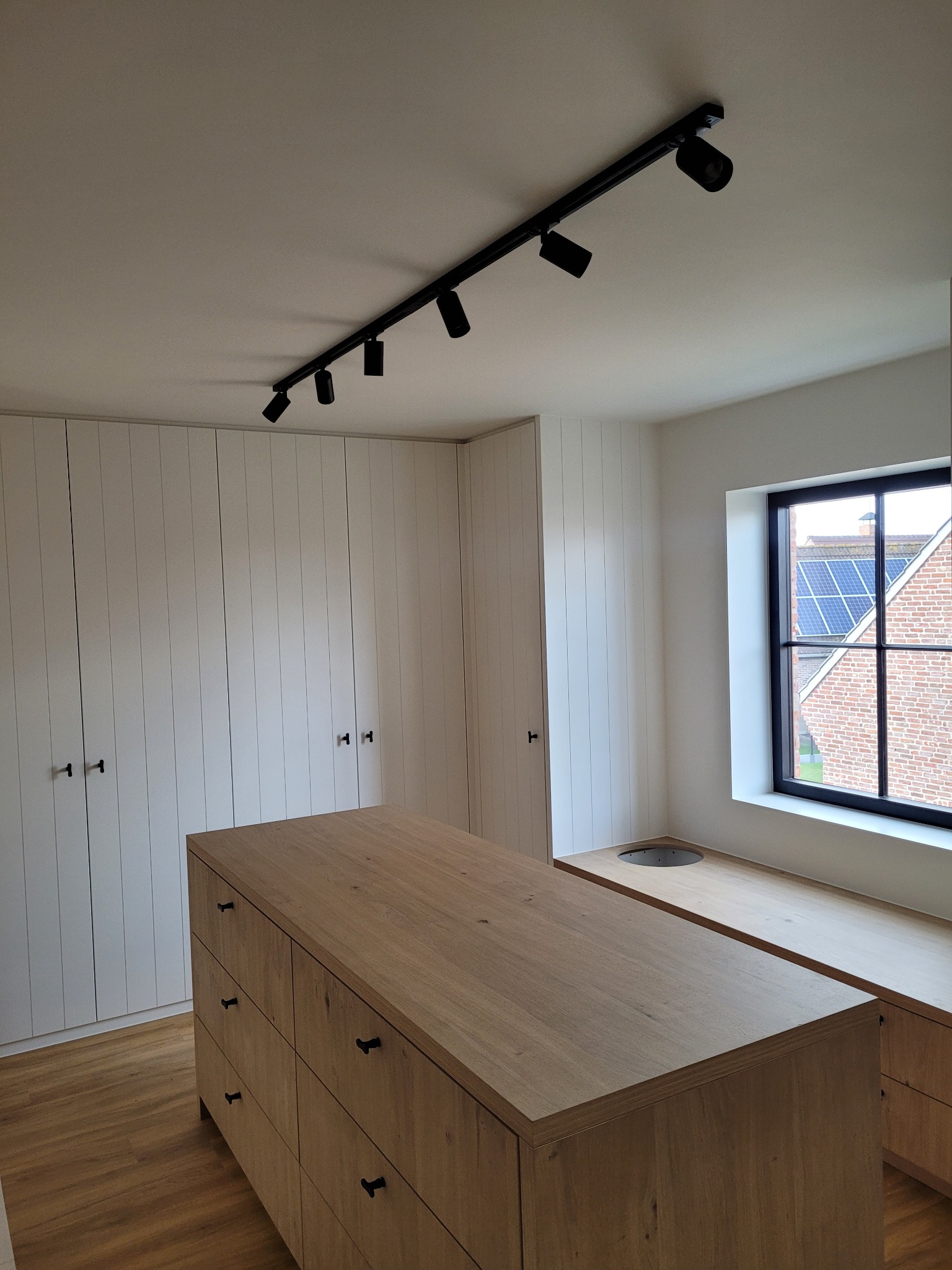 Czarna szyna z reflektorami sufitowymi oświetlająca drewnianą wyspę z szufladami w nowoczesnej garderobie z białymi szafami w zabudowie i oknem z widokiem na dom z panelami słonecznymi.