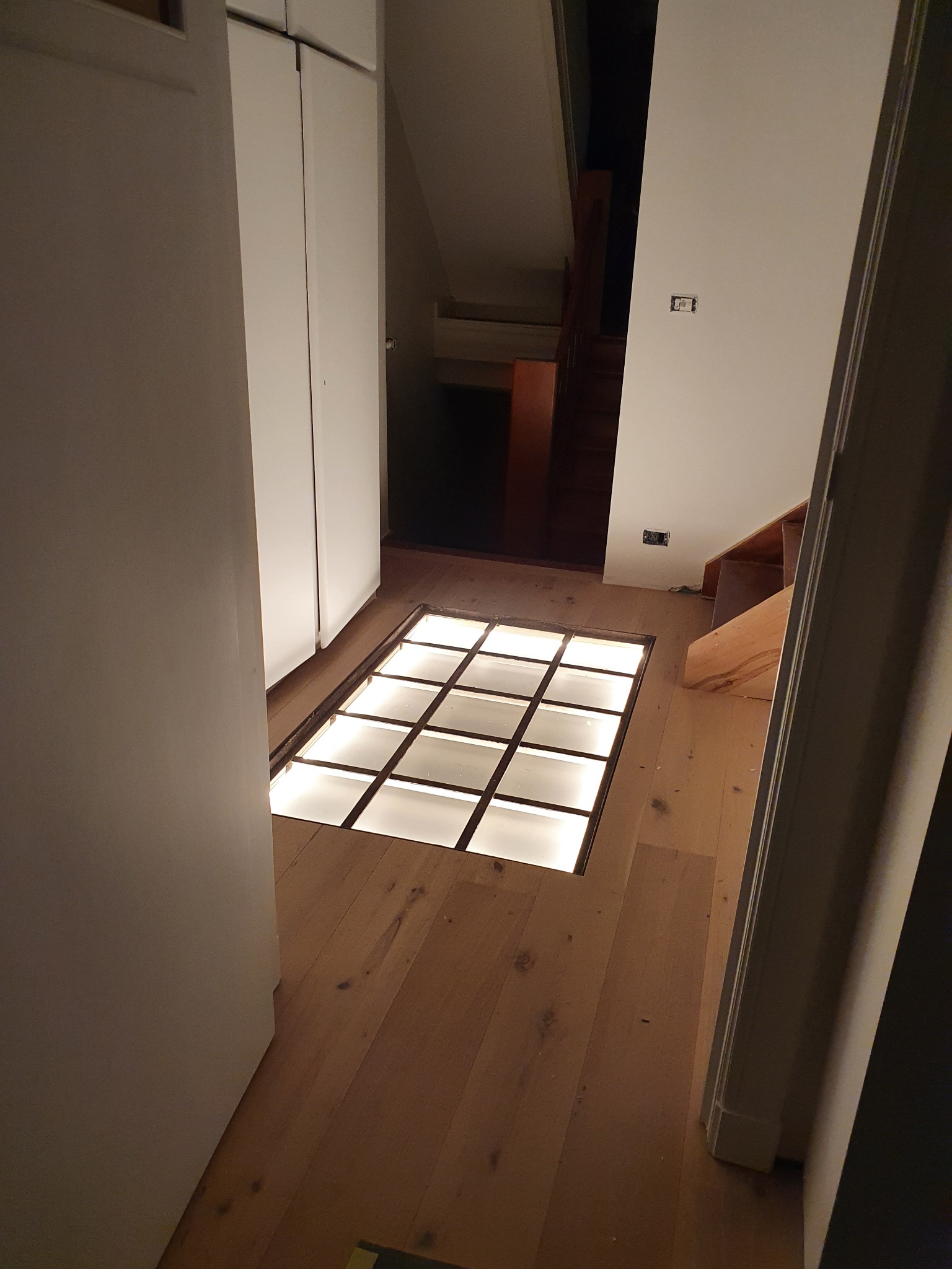 Podświetlana szklana podłoga w korytarzu z drewnianą podłogą i białymi szafami w tle.