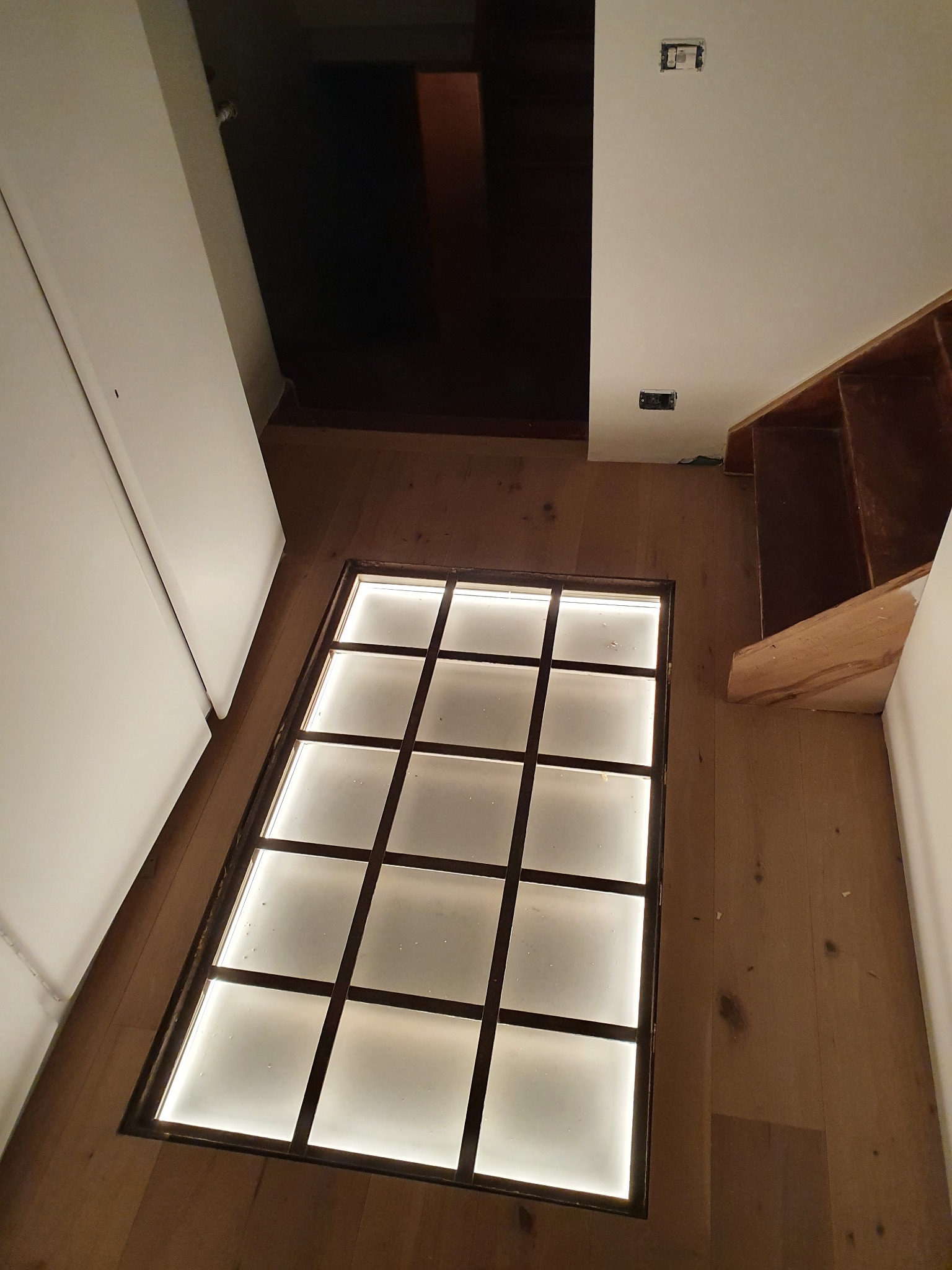 Podświetlana podłoga z kwadratowymi panelami wkomponowana w drewnianą podłogę, widok z góry na korytarz z elementami w trakcie wykończenia.