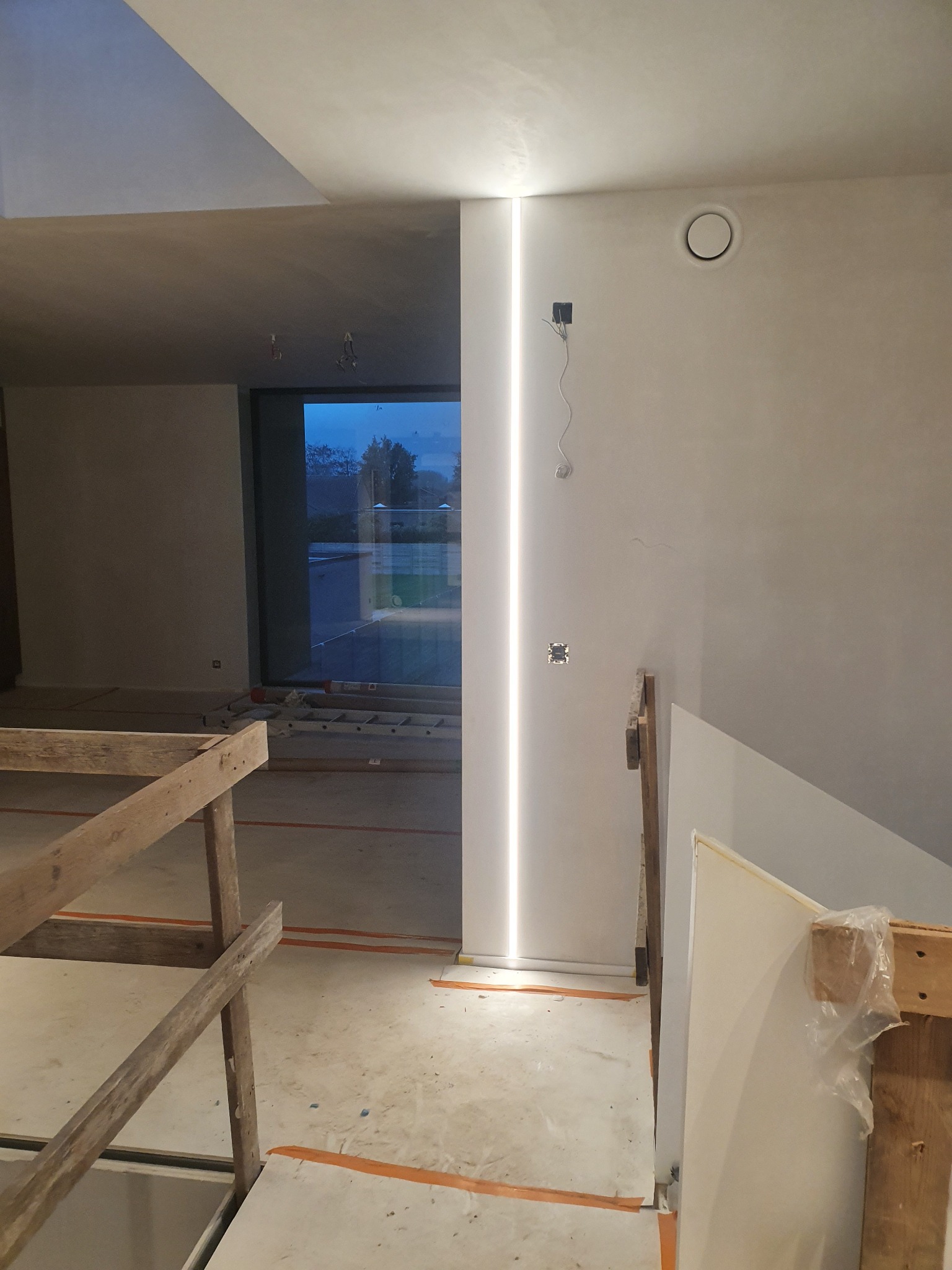 Wnętrze w trakcie wykańczania z pionową listwą LED w ścianie, widoczne okno z widokiem na zewnątrz, fragment rusztowania i wystające kable elektryczne.