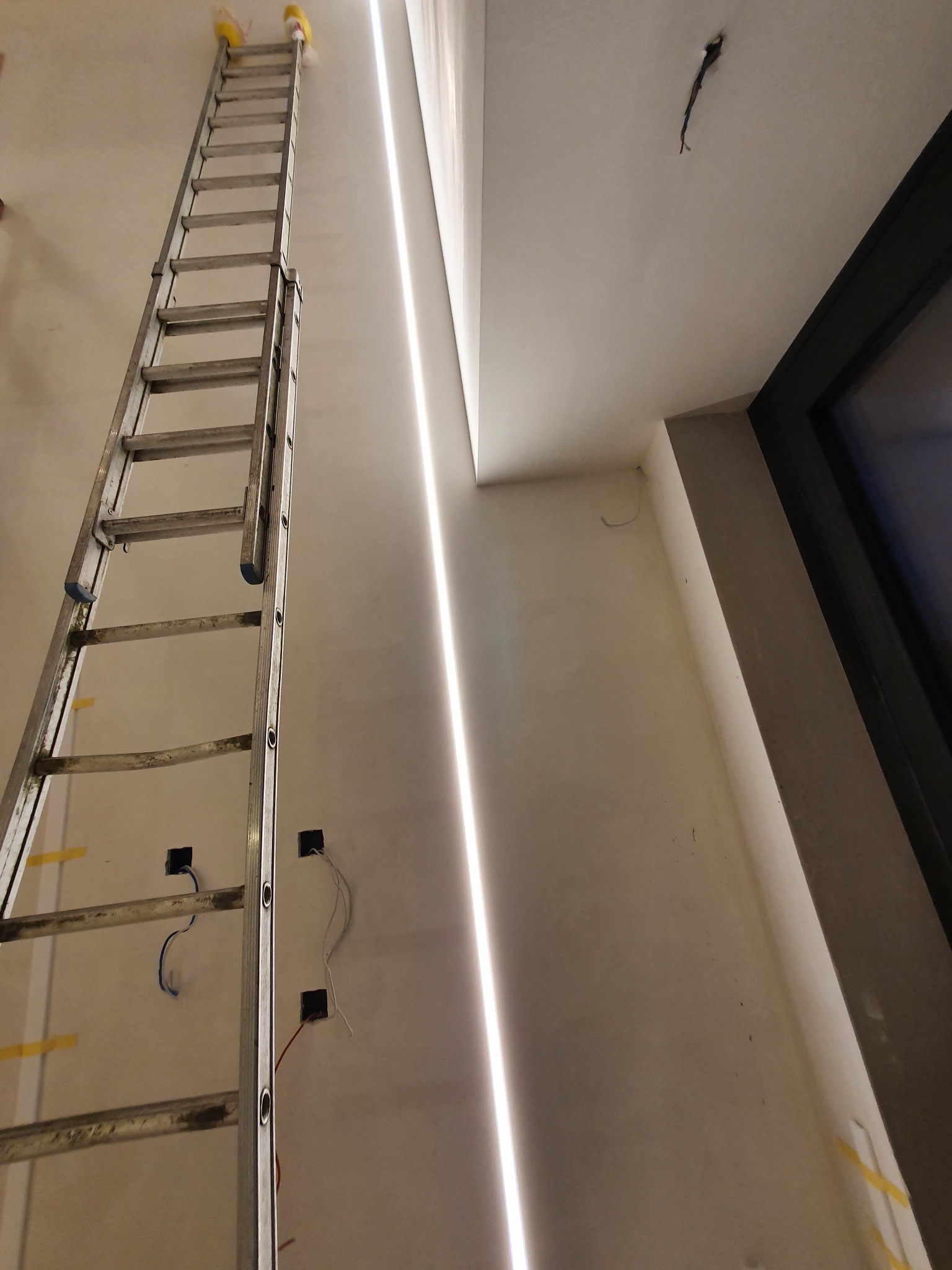 Wysoka aluminiowa drabina oparta o ścianę z zamontowanym liniowym oświetleniem LED. Widoczne puszki elektryczne z wystającymi przewodami oraz otwór w suficie z kablami.