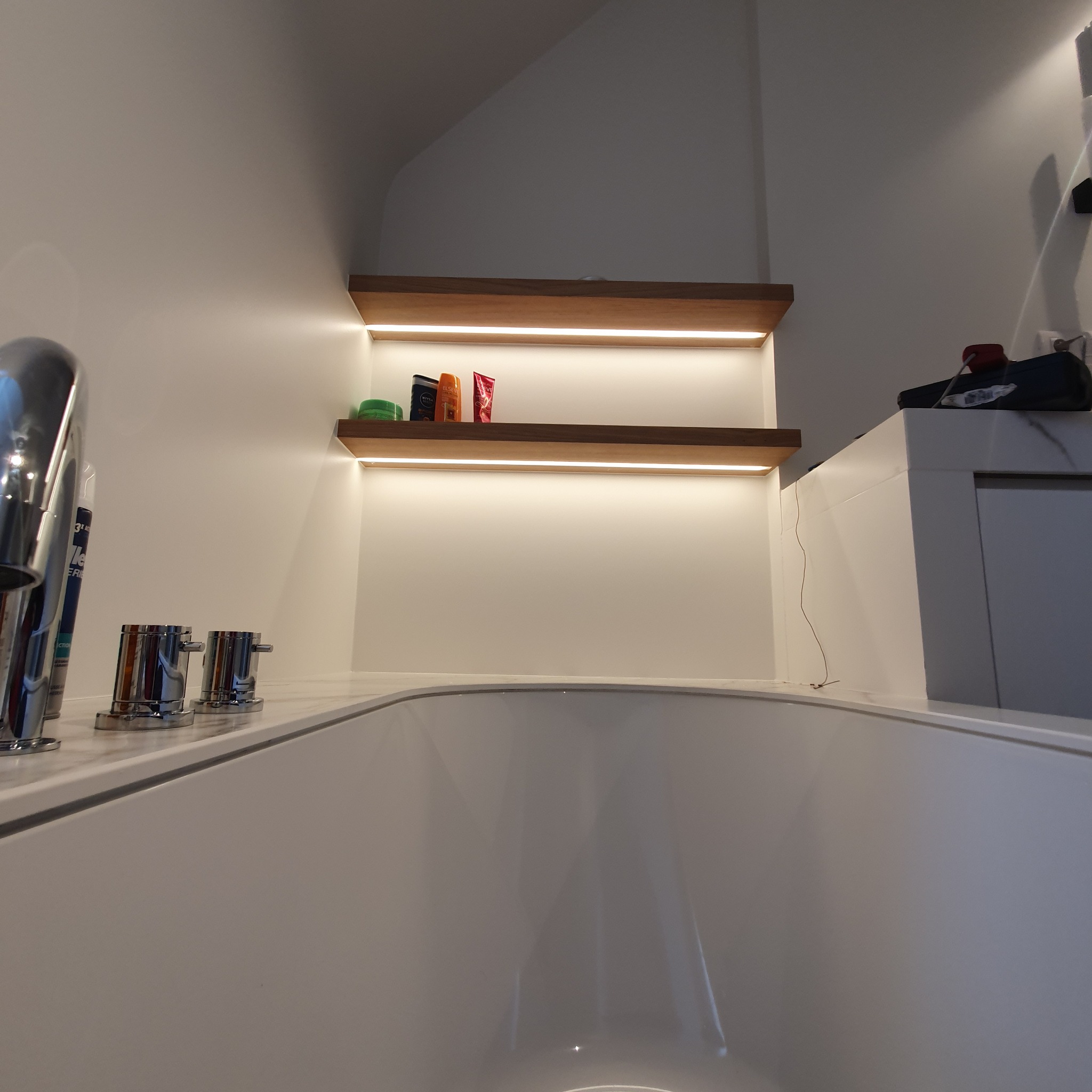 Nowoczesna łazienka z podświetlanymi półkami, widok z perspektywy wanny, chromowana bateria umywalkowa, kosmetyki na półkach.