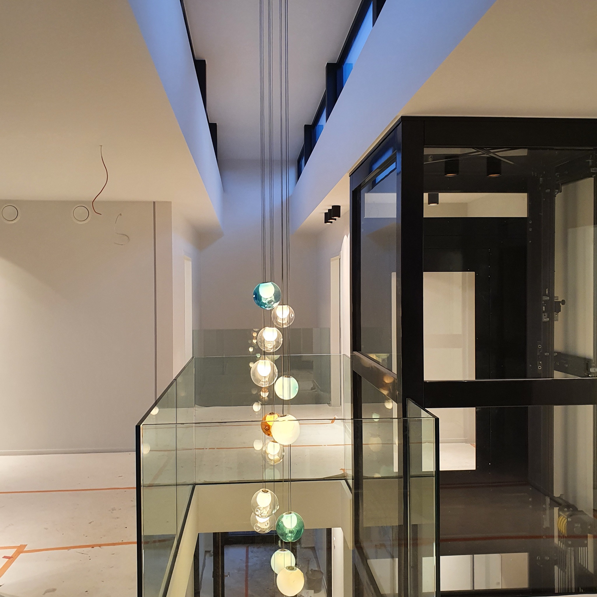 Nowoczesny hol z szklaną balustradą, widok na dół, w tle winda osobowa i wisząca lampa składająca się z wielu szklanych kul w różnych kolorach, na ścianie widoczne przewody elektryczne.