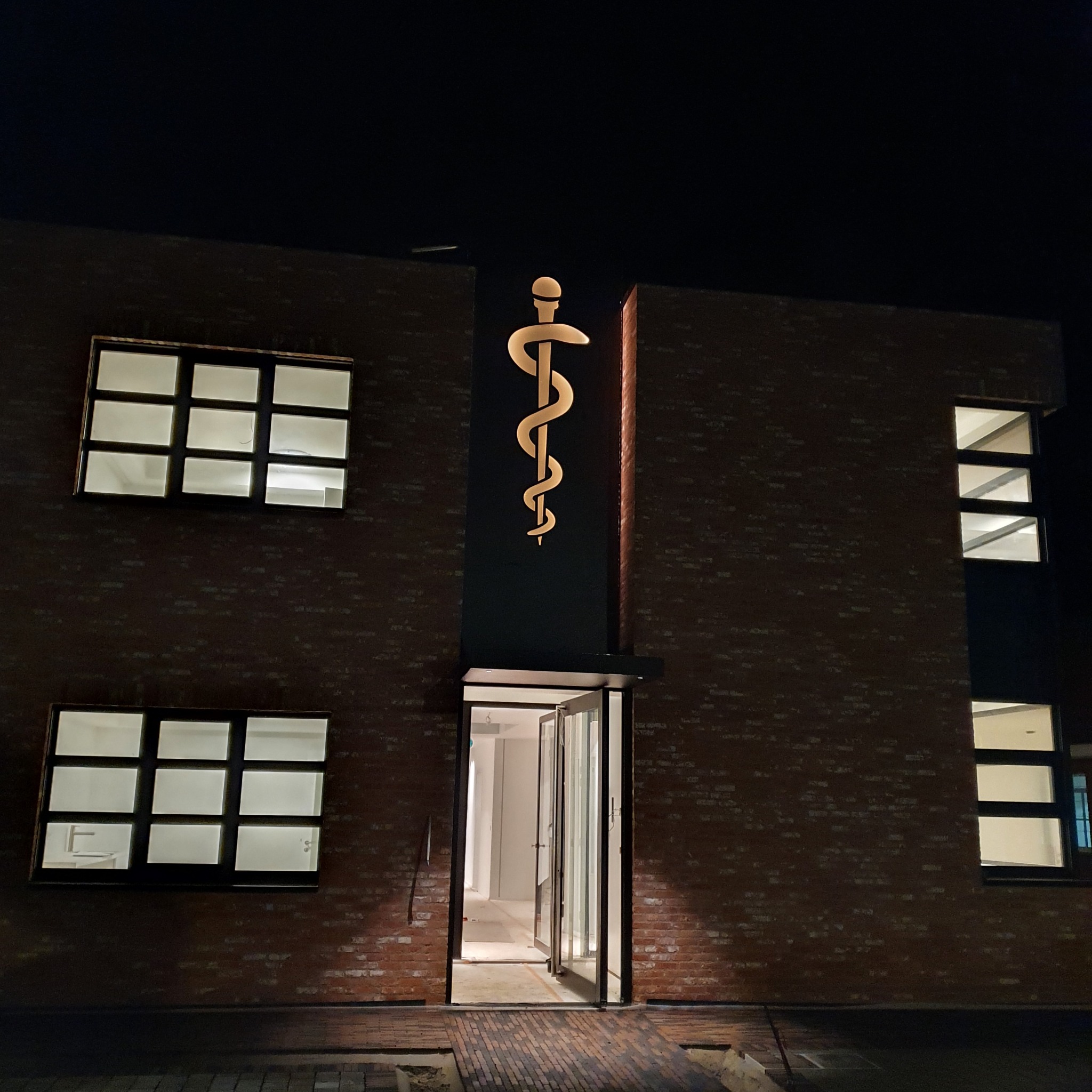 Budynek z cegły z podświetlonym symbolem Eskulapa nad otwartymi drzwiami wejściowymi, widoczne okna z białym światłem wewnątrz.