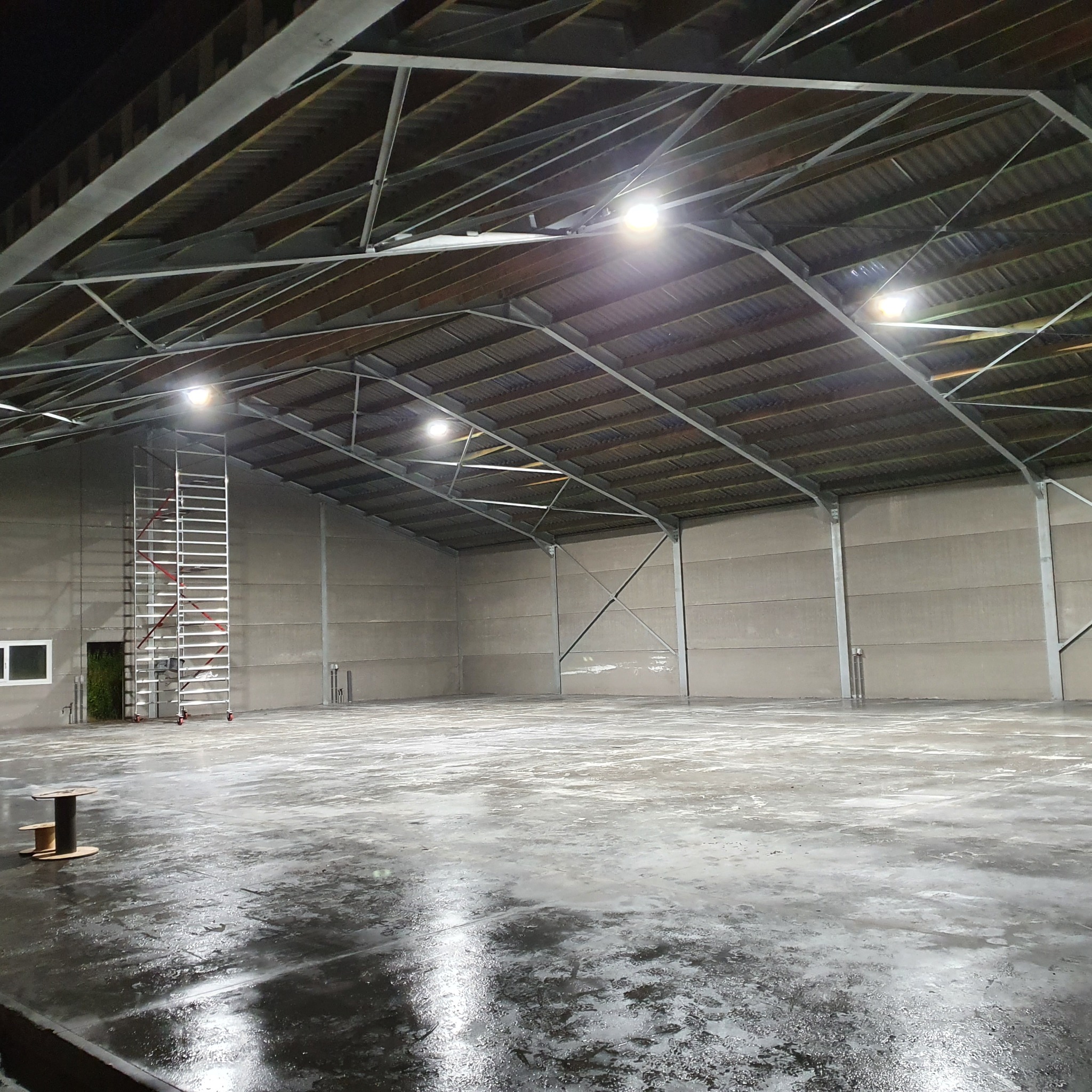 Wnętrze pustego magazynu z betonową podłogą, oświetleniem LED pod sufitem i drabiną na kółkach przy ścianie. Na podłodze widoczna szpula z kablem.