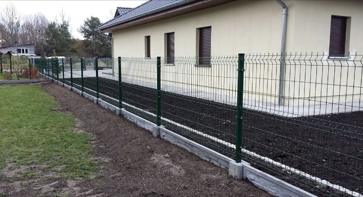 Zielone ogrodzenie panelowe z podmurówką betonową, widok z zewnątrz posesji, świeżo obsypana ziemia po obu stronach ogrodzenia.