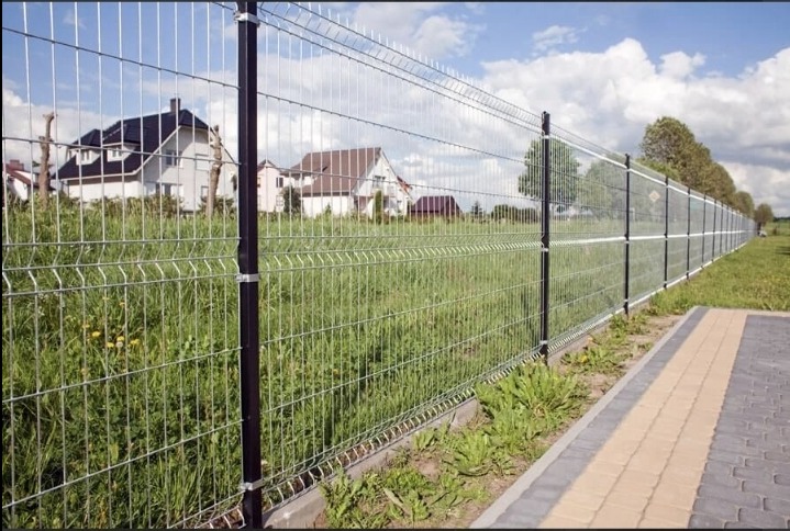 Długi, metalowy płot panelowy z czarnymi słupkami, widok z poziomu chodnika z kostki brukowej, w tle domy jednorodzinne i zielona łąka.
